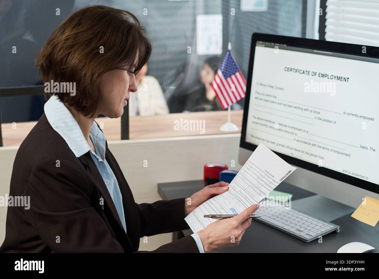 Donna caucasica di mezza età che esamina i documenti alla scrivania del centro visti, tiene i documenti e si siede di fronte al monitor del computer e mostra il modulo del certificato di impiego Foto Stock