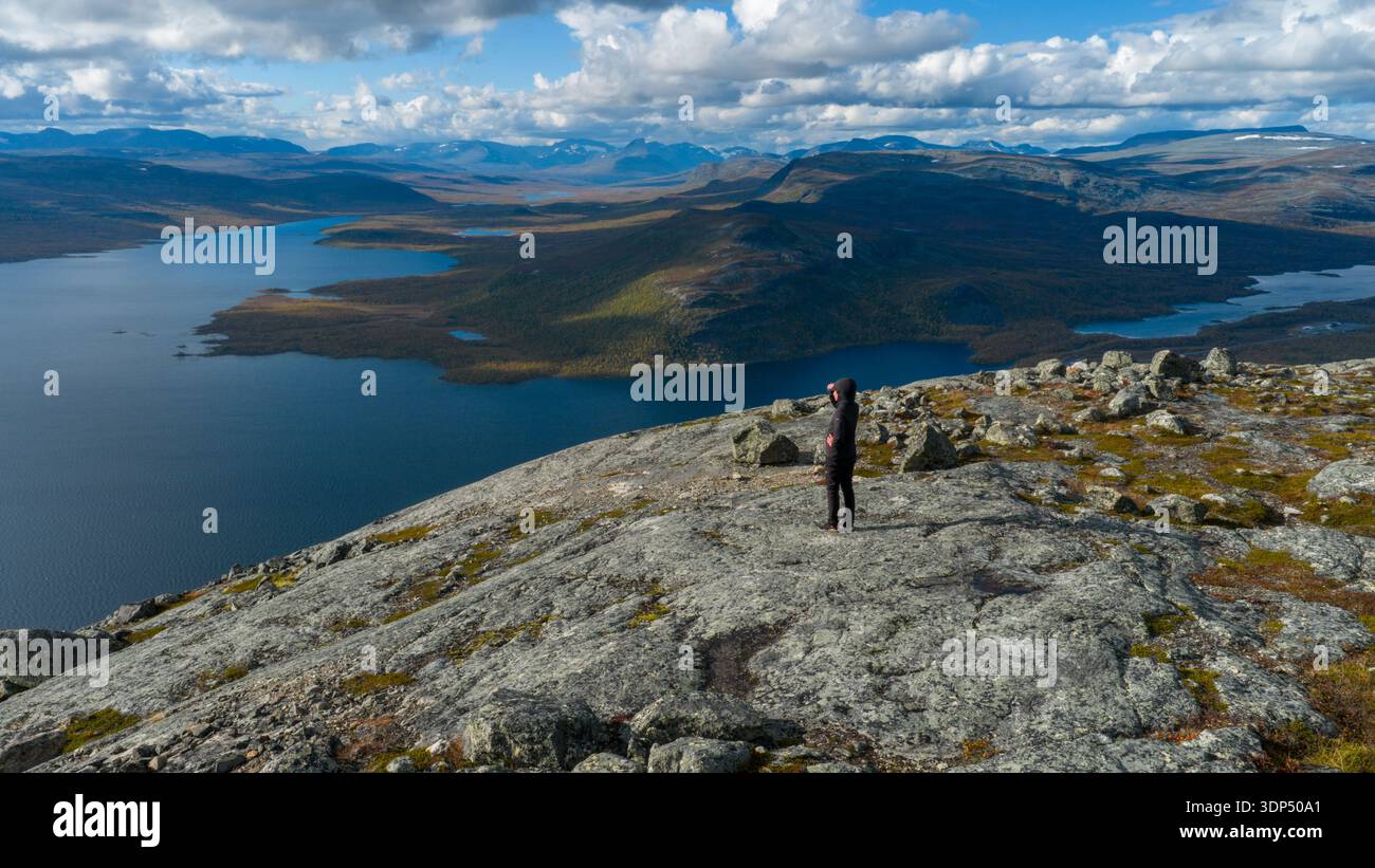 La vista aerea di una figura solitaria si erge in cima a una cima rocciosa che si affaccia sulle vaste e tranquille acque di un lago circondato da aspre montagne in lontananza, Kil Foto Stock