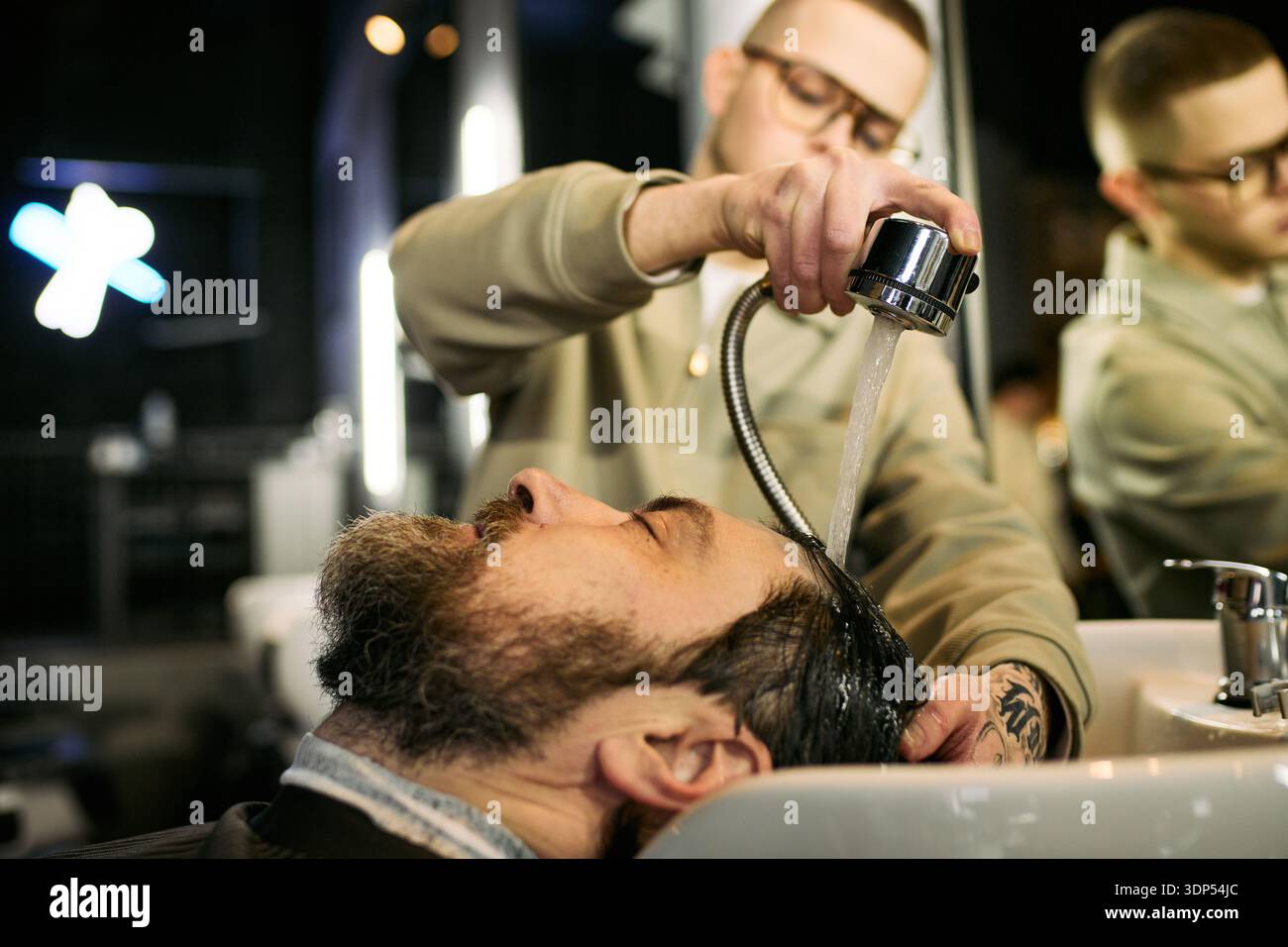 Giovane uomo adulto caucasico che lava i capelli di un uomo caucasico di mezza età al lavandino del barbiere, concentrandosi sul processo di lavaggio professionale dei capelli con acqua che scorre sopra la testa dei clienti Foto Stock
