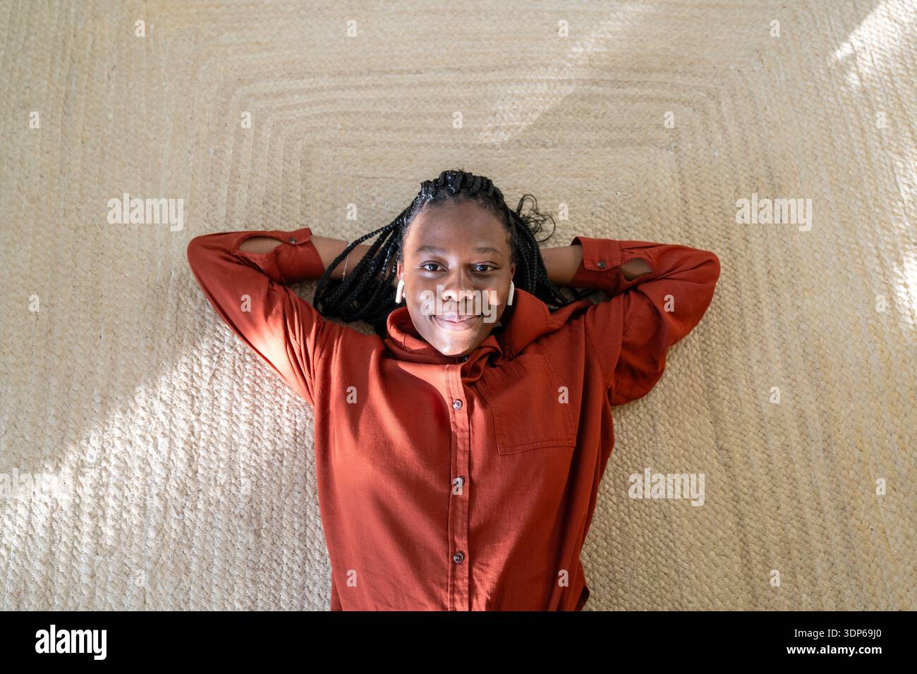 Donna afroamericana ottimista e allegra sdraiata su un tappeto di iuta a casa ad ascoltare musica in baccelli. Foto Stock