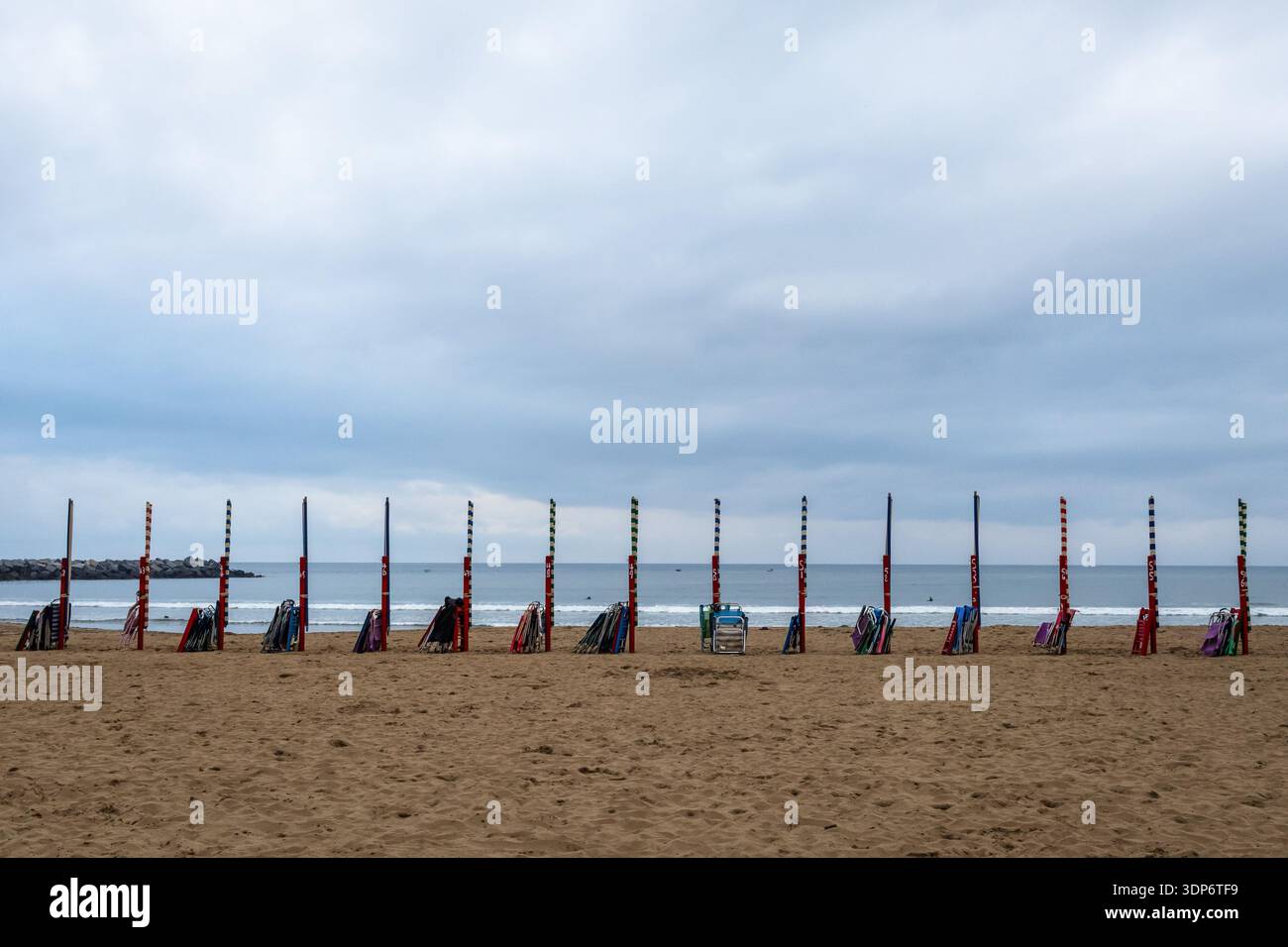 Una lunga e simmetrica fila di ombrelloni chiusi e pali di legno allineati su una spiaggia sabbiosa sotto un cielo nuvoloso. Foto Stock