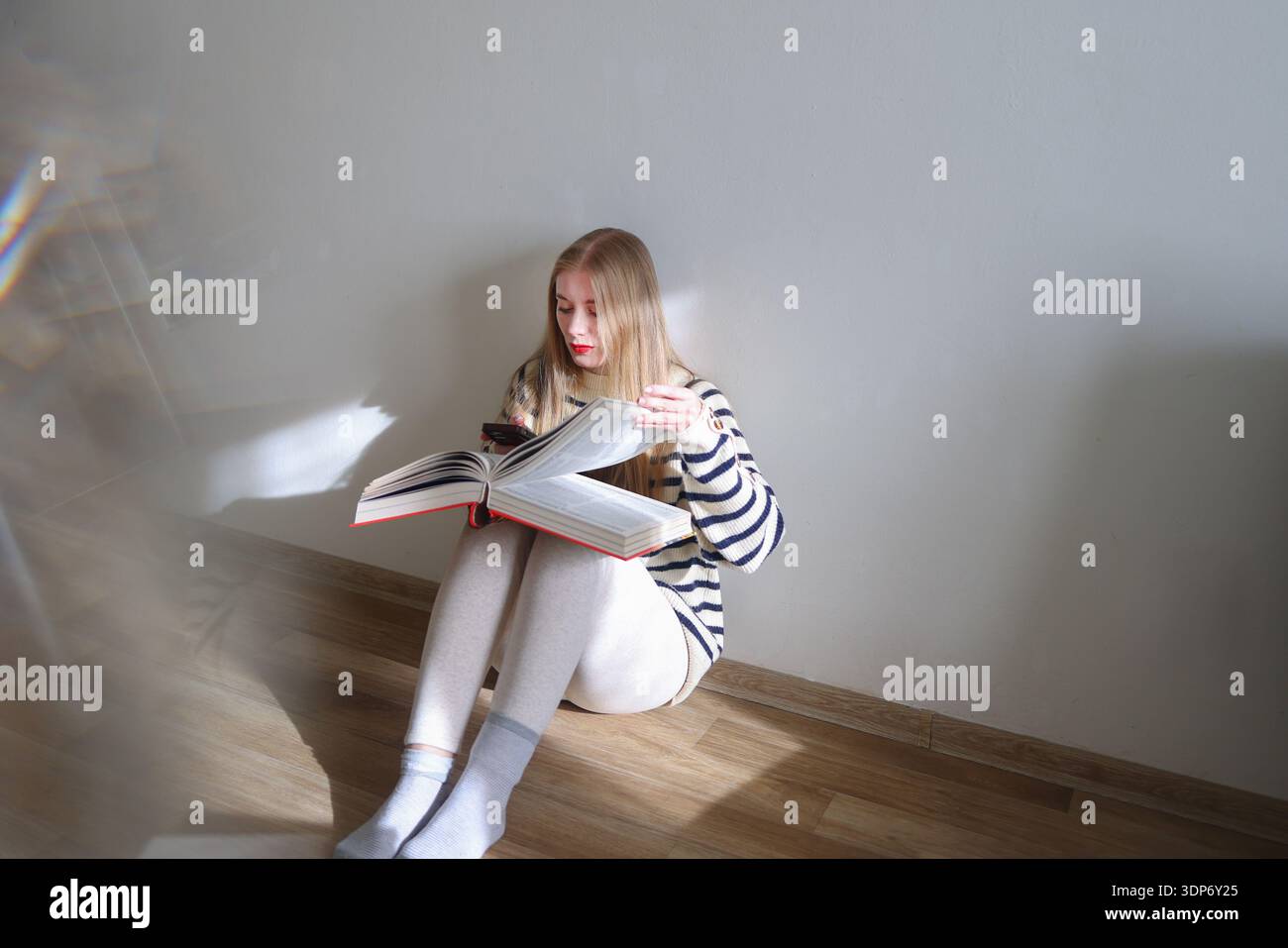 Giovane donna seduta su pavimento di legno e che legge un grande libro aperto a casa. Stile di vita accogliente, educazione e auto-sviluppo con spazio di copia Foto Stock