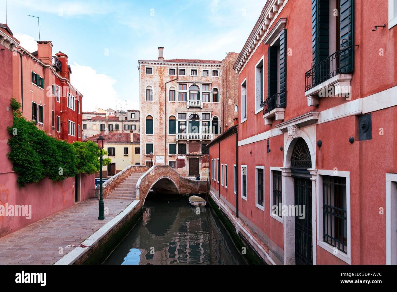 Un tranquillo canale veneziano con un ponte ad arco, edifici in terracotta e una passerella acciottolata sotto il cielo blu. Foto Stock