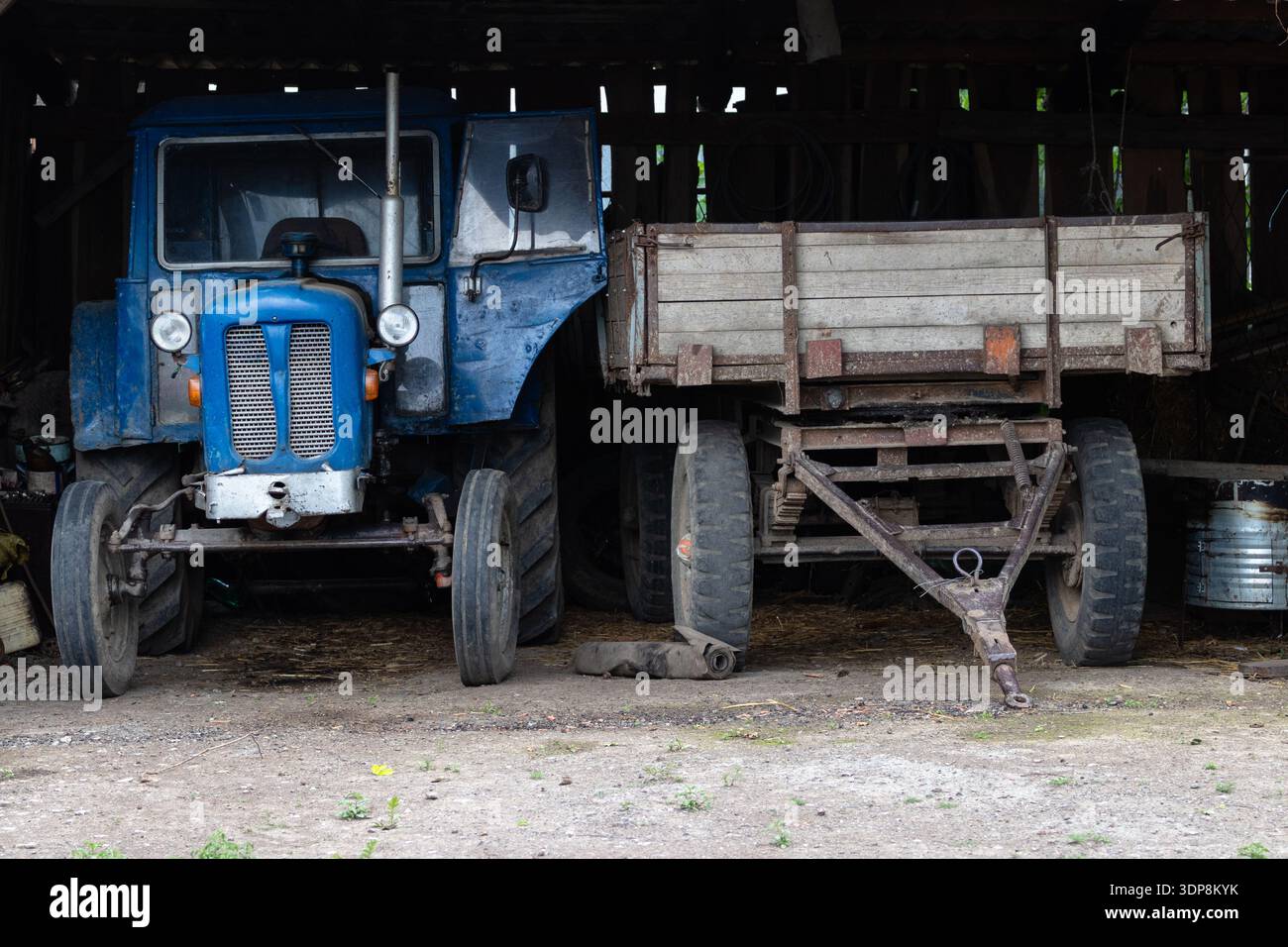 Un vecchio trattore blu abbinato a un rimorchio in legno intempestivo parcheggiato all'interno di un fienile rustico, che evoca l'agricoltura rurale, le attrezzature agricole d'epoca, il "tex" Foto Stock