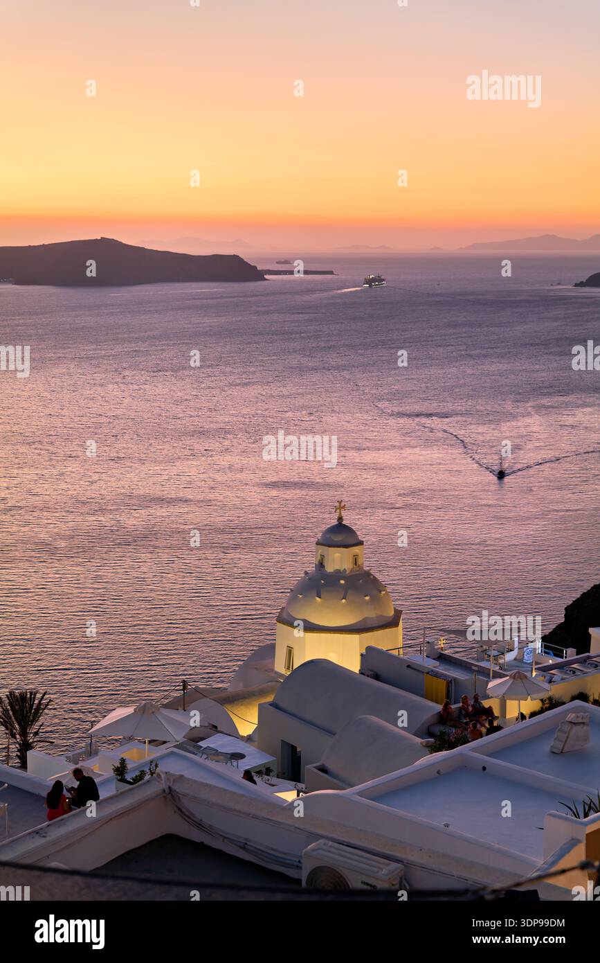 Un tranquillo tramonto a Santorini, in Grecia, illumina una cupola della chiesa e una caldera, mentre le persone si rilassano sui tetti e le barche navigano sul Mar Egeo. Foto Stock