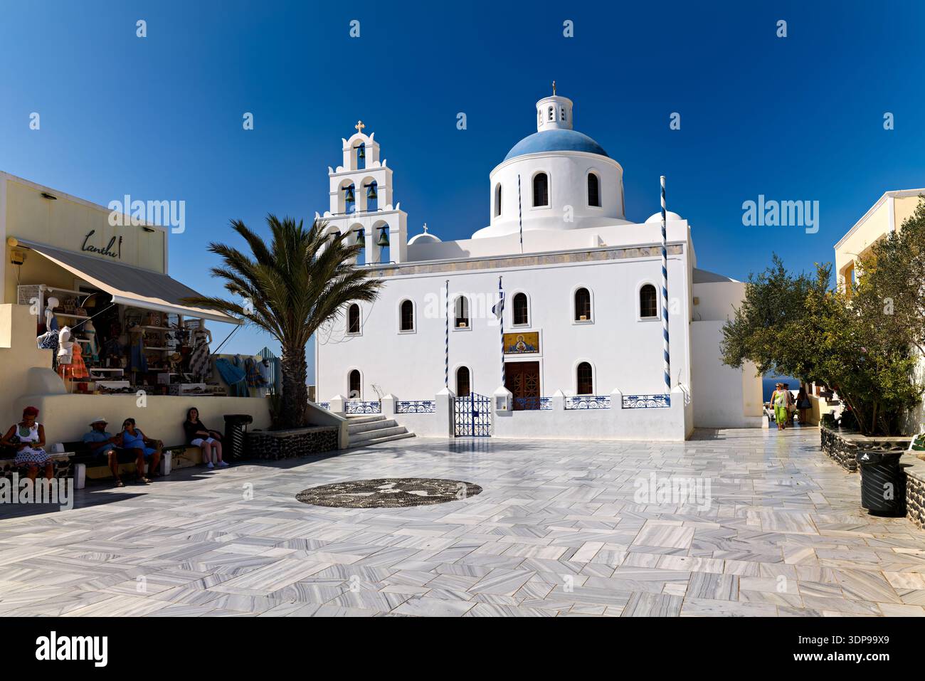 I turisti apprezzano la soleggiata piazza fuori dalla chiesa di Panagia Platsani a Oia, Santorini, Grecia, ammirando l'iconica architettura bianca e la cupola blu. Foto Stock