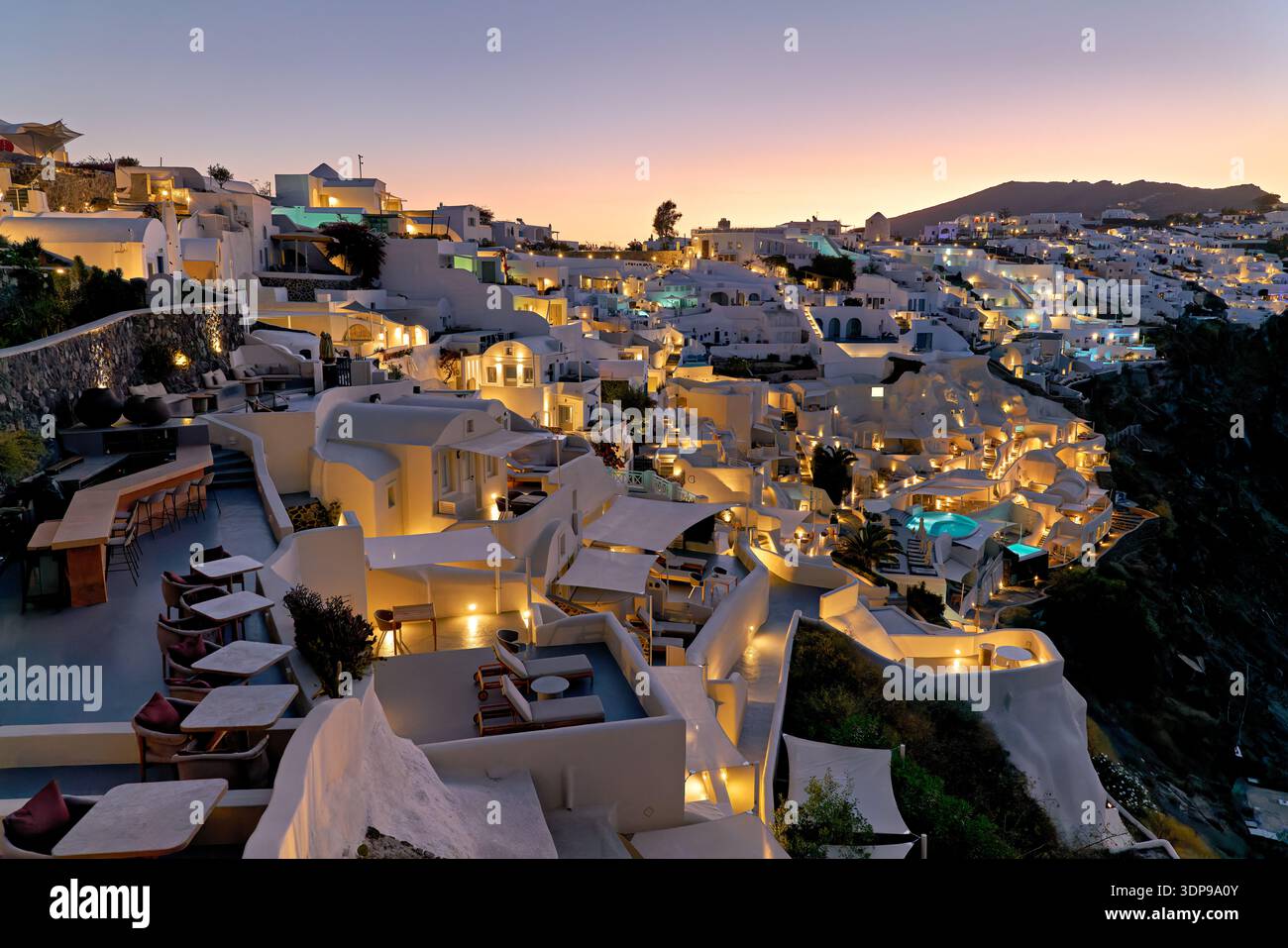 Oia, Santorini, Grecia, al crepuscolo. Gli edifici imbiancati di bianco e gli hotel brillano di luci, offrendo ai visitatori viste mozzafiato che si godono l'atmosfera serale Foto Stock