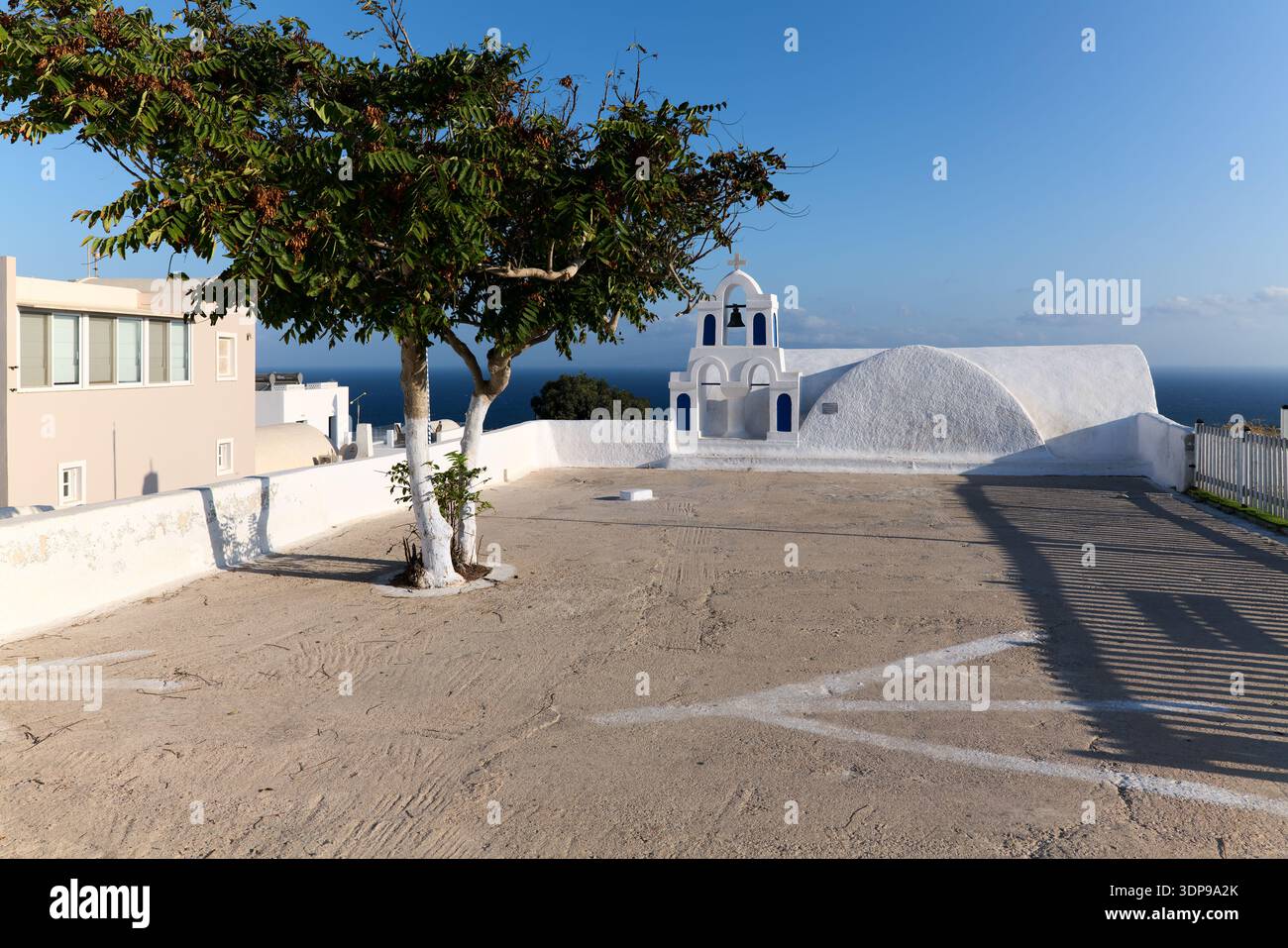 Una tradizionale chiesa bianca con un campanile blu a Santorini, in Grecia, si affaccia sul Mar Egeo. Un albero si trova in primo piano, catturando l'islan Foto Stock