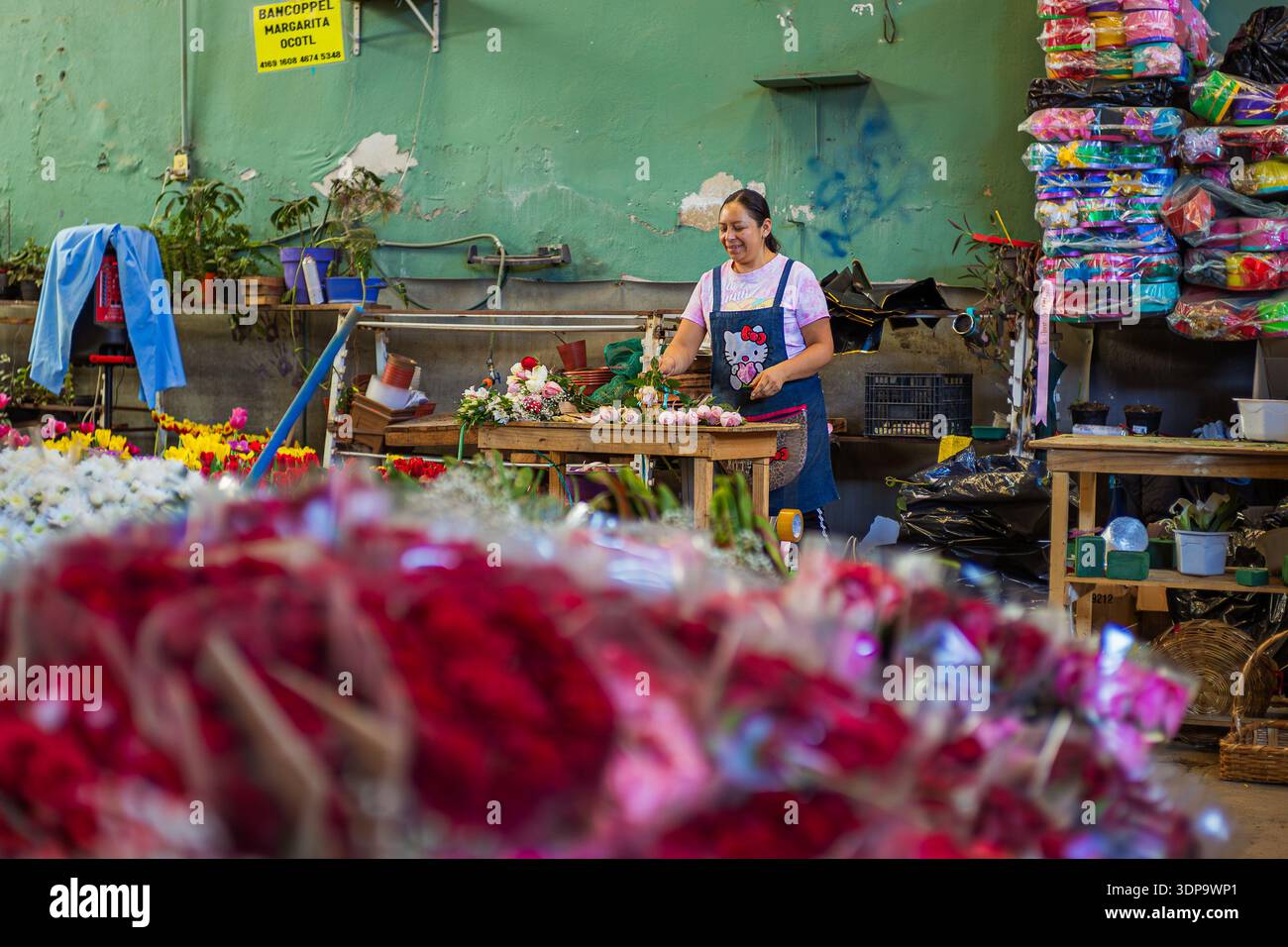 Una donna sorridente organizza composizioni floreali in un tradizionale bancone del mercato, circondato da rose rosse e forniture floreali. Foto Stock