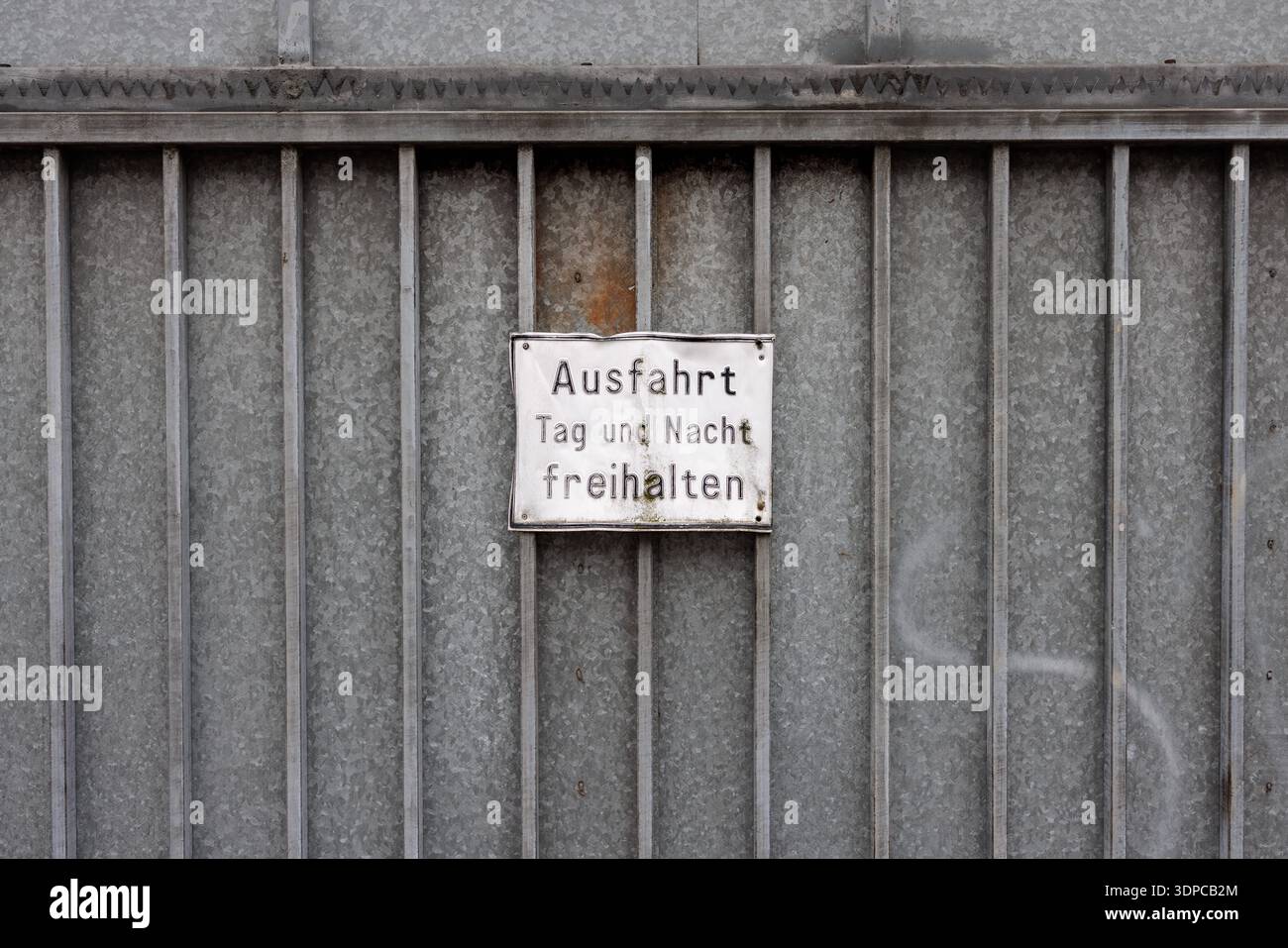Ausfahrt Tag und Nacht freihalten - Ein weißes, leicht verwittertes und verbogenes Hinweisschild mit dem Hinweis *Ausfahrt Tag und Nacht freihalten* hängt am 13.02.2026 an einem verzinkten Metalltor in Osnabrück Niedersachsen, Deutschland. Solche Schilder dienen im städtischen Raum als präventiver Hinweis auf privatrechtliche Parkverbote und markieren kritische Zufahrtswege für Rettungskräfte oder Anwohner. *** Tenere l'uscita limpida giorno e notte Un cartello bianco, leggermente intemprato e piegato con il messaggio Ausfahrt Tag und Nacht freihalten è appeso su un cancello metallico galvanizzato a Osnabrück bassa Sassonia, GE Foto Stock