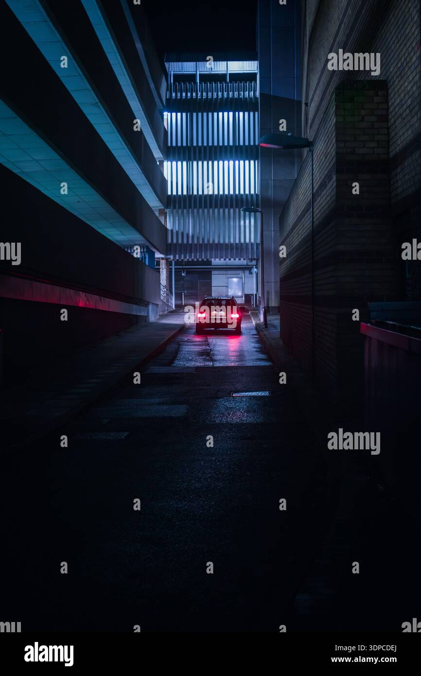 Auto con luci di arresto illuminate che attraversa un vicolo urbano contemporaneo incorniciato da edifici moderni con illuminazione a LED verticale al crepuscolo Foto Stock