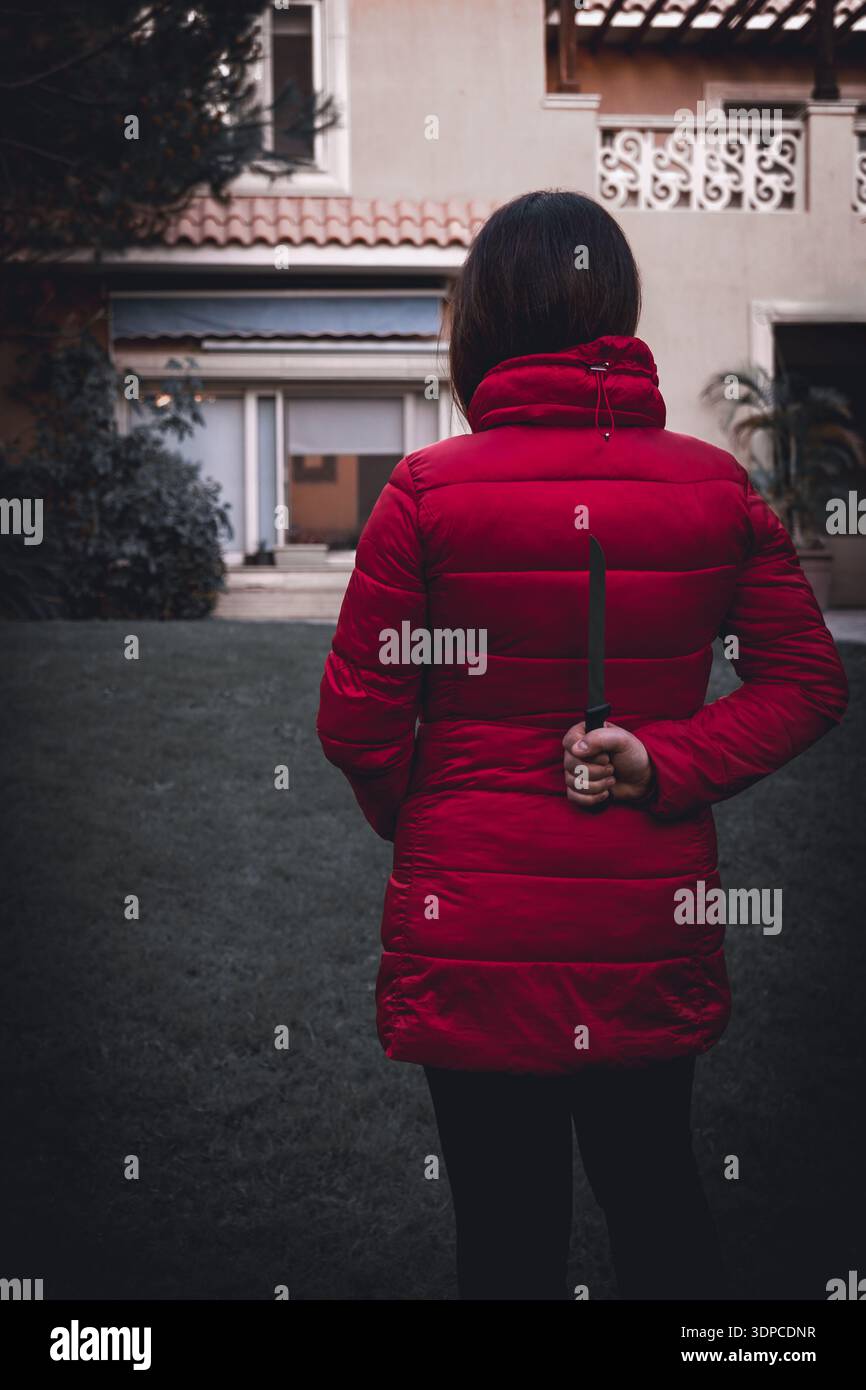 Donna in giacca rossa che tiene il coltello dietro l'edificio residenziale di fronte all'edificio in un thriller concettuale o in un'immagine di suspense Foto Stock