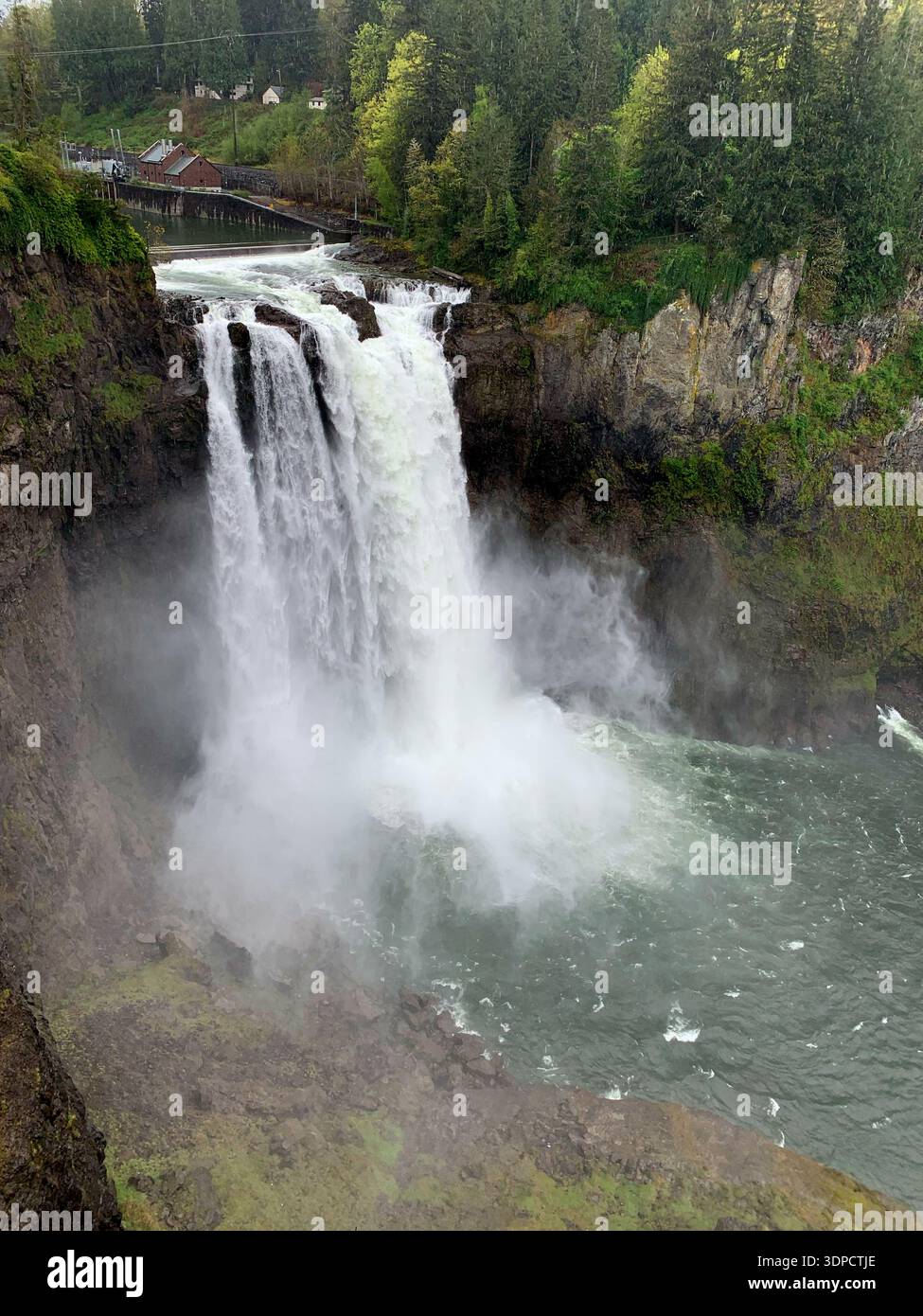 Maestosa cascata che scende giù dalle scogliere rocciose circondate da lussureggiante vegetazione e nebbia, con un ponte in legno e edifici visibili sullo sfondo Foto Stock