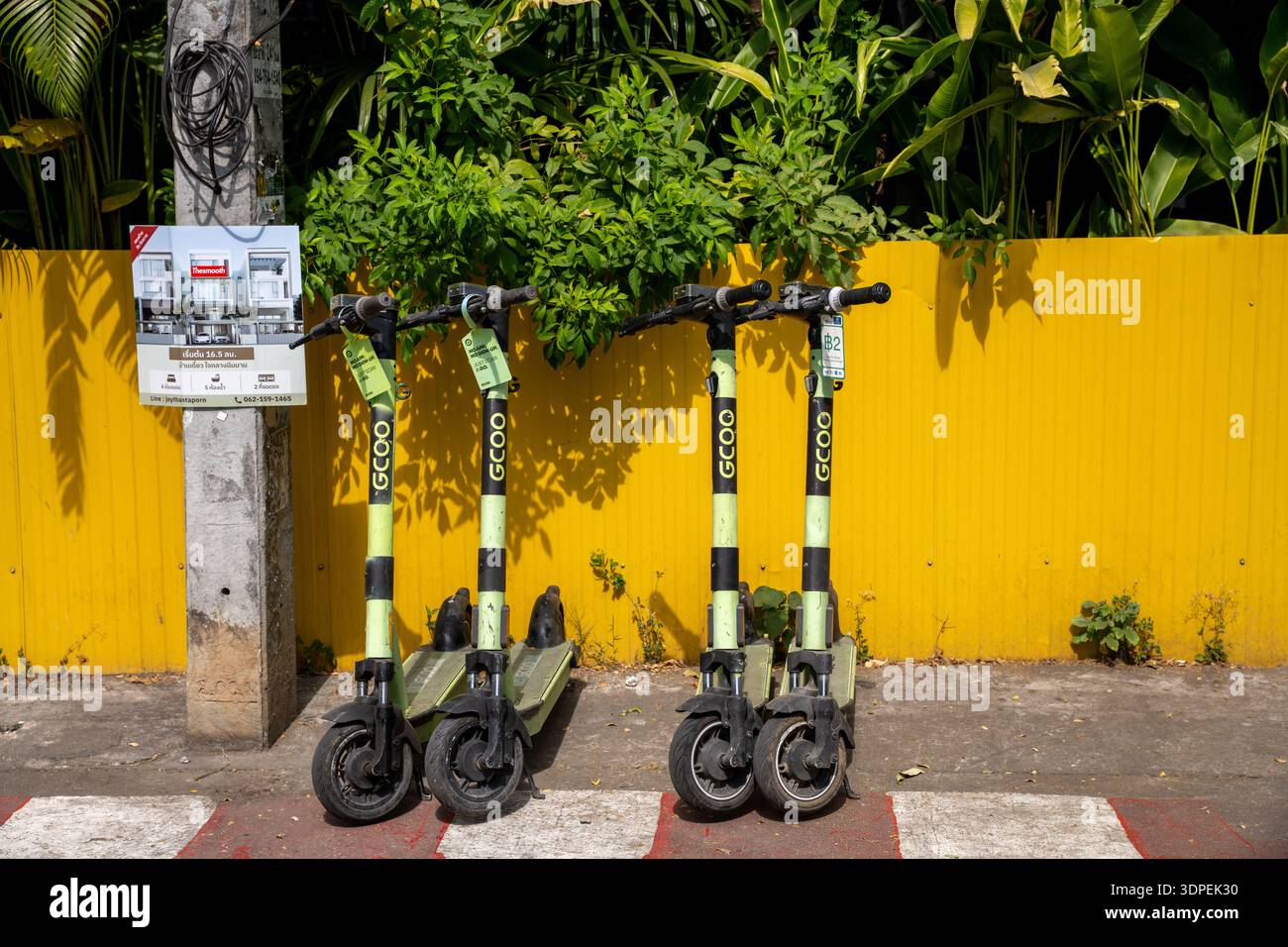 Una linea di scooter elettrici GCOO disponibili per il noleggio pubblico su un marciapiede in Thailandia. La scena è ambientata contro una recinzione corrugata di colore giallo Foto Stock