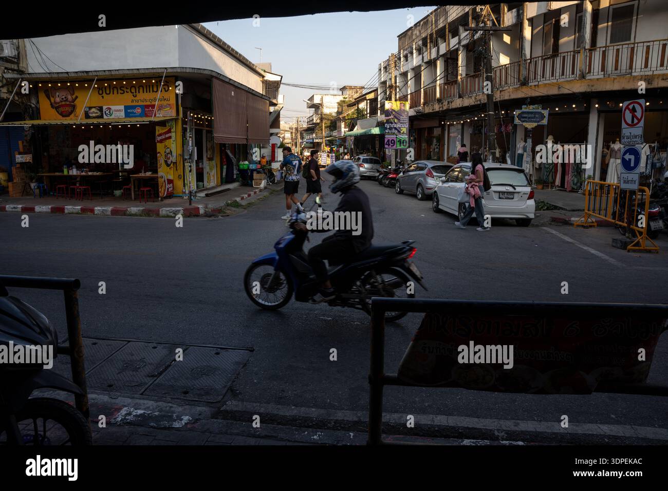Una persona che indossa un casco guida uno scooter attraverso un incrocio stradale affollato in una città della Thailandia. Il caldo sole del tardo pomeriggio illumina il mestiere Foto Stock