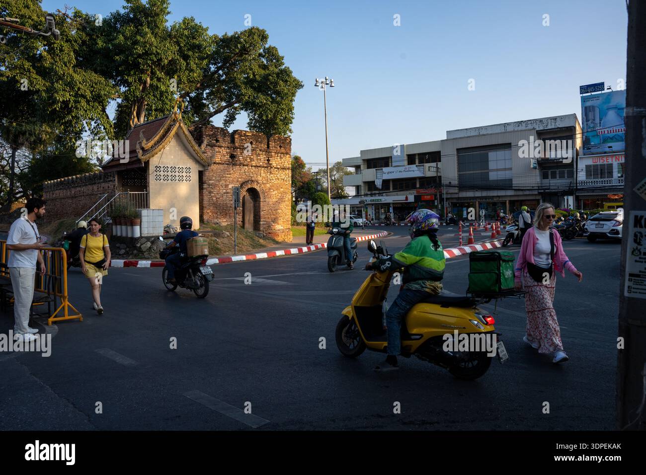 Un vivace angolo di strada a Chiang mai, Thailandia, durante un pomeriggio soleggiato. I turisti e la gente del posto navigano nel traffico di scooter vicino a una storica ga di mattoni Foto Stock