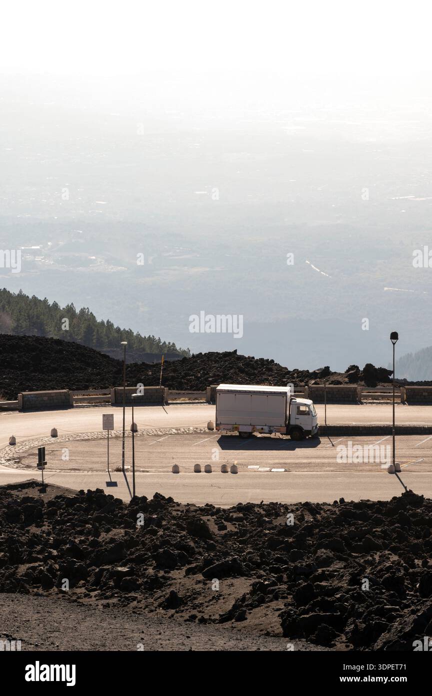 Un camion bianco solitario è parcheggiato in un'area pavimentata quasi vuota sulle pendici vulcaniche dell'Etna. Rocce di lava scure e frastagliate dominano il primo piano, wh Foto Stock