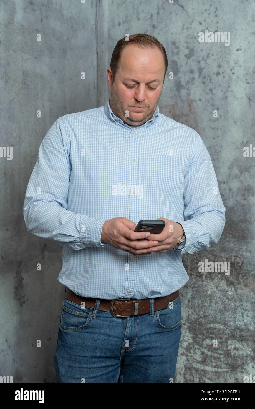 Uomo con camicia blu concentrato sullo schermo del telefono in interni. Concetto di comunicazione moderno, lavoro in remoto, SMS, navigazione in Internet e uso quotidiano della tecnologia Foto Stock