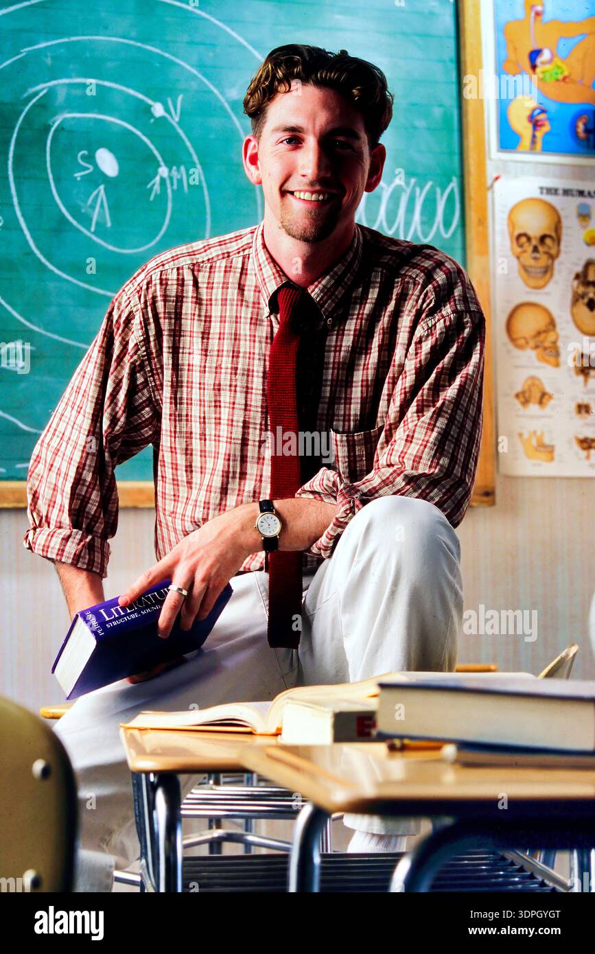 Un ritratto di un insegnante di sesso maschile in piedi su una scrivania in una classe. Tiene un libro e sorride mentre è circondato da carte didattiche e da una lavagna. St Foto Stock