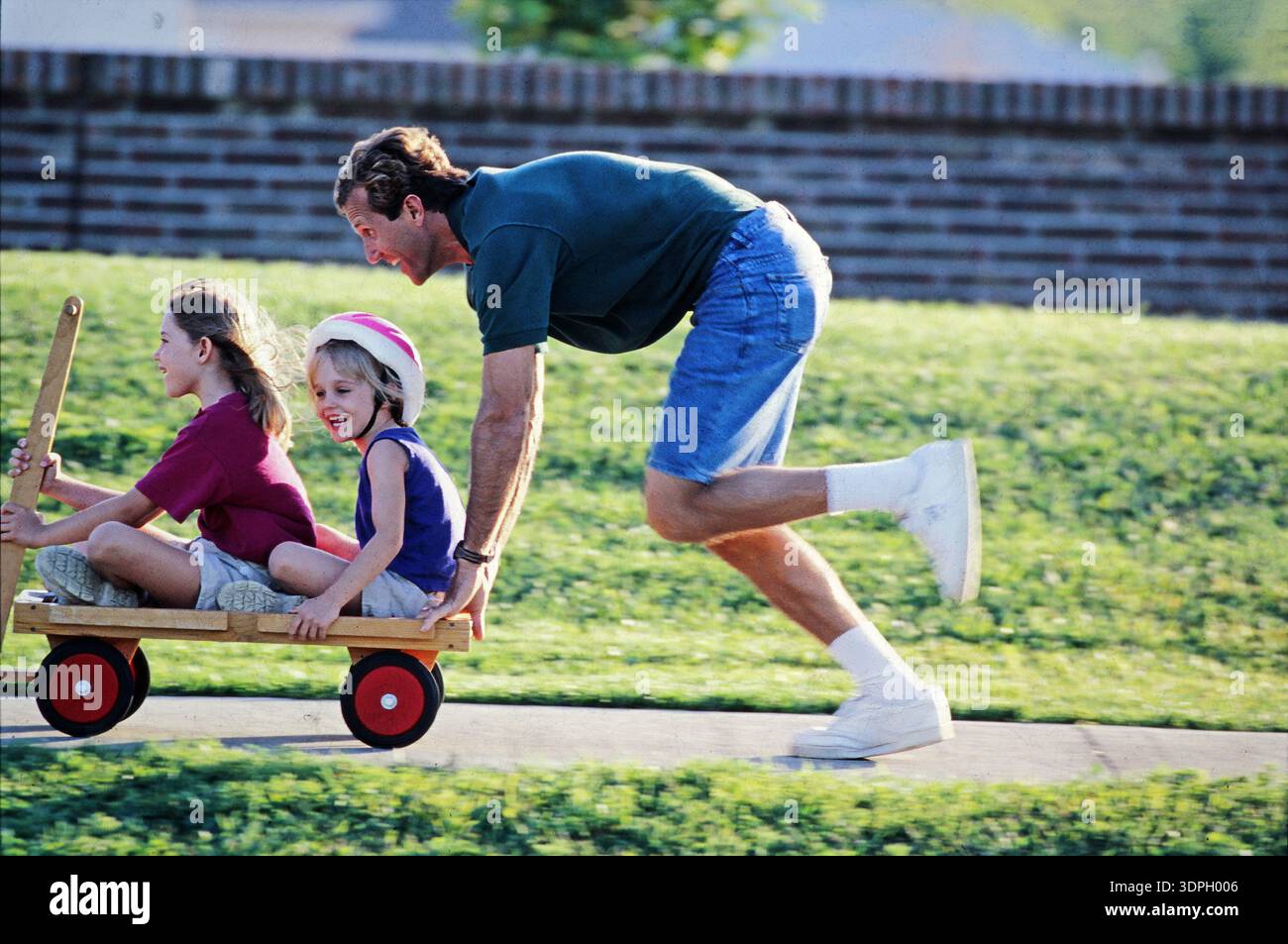 Un padre spinge un carro con i suoi due figli seduti dentro. I bambini sorridono mentre si divertono. L'ambiente è un cortile verde in un vicino Foto Stock