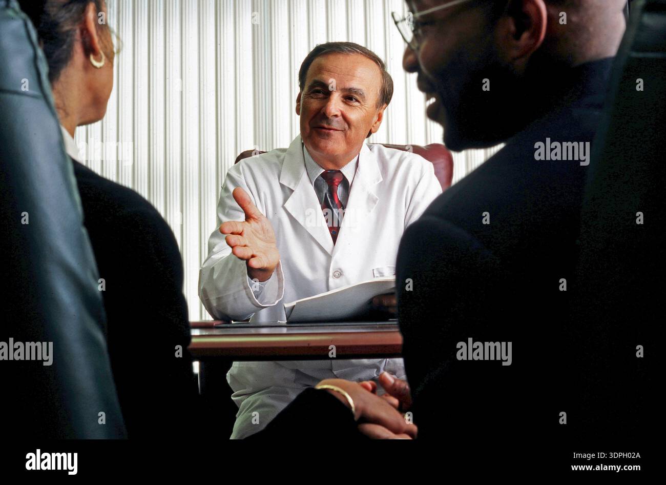 Un medico con un cappotto bianco è seduto ad una scrivania e parla con una giovane coppia. Sembrano essere impegnati in una conversazione sulla salute e il trattamento o Foto Stock