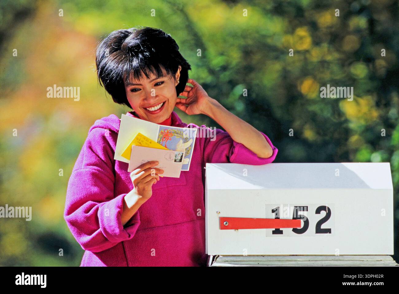 E l'attraente donna asiatica sorride mentre si trova accanto a una cassetta postale che legge la sua posta. Ha in mano diverse cartoline e lettere. Il Suor Foto Stock