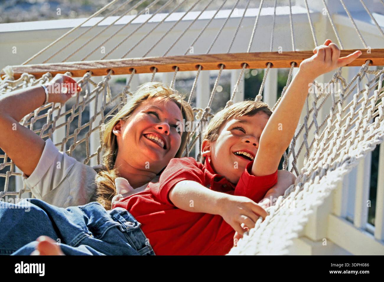 Una madre e il suo giovane figlio si rilassano in un'amaca, ridendo e giocando. La scena mostra i loro momenti di gioia, con la luce del sole che splende su di loro e una luce brillante Foto Stock