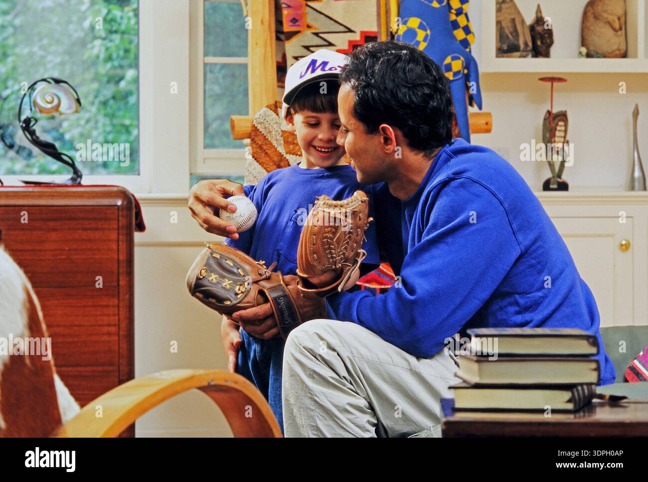 Un padre e un figlio latino si divertono insieme mentre guardano un baseball in salotto. Il padre sorride al figlio, che tiene in mano un guanto Foto Stock