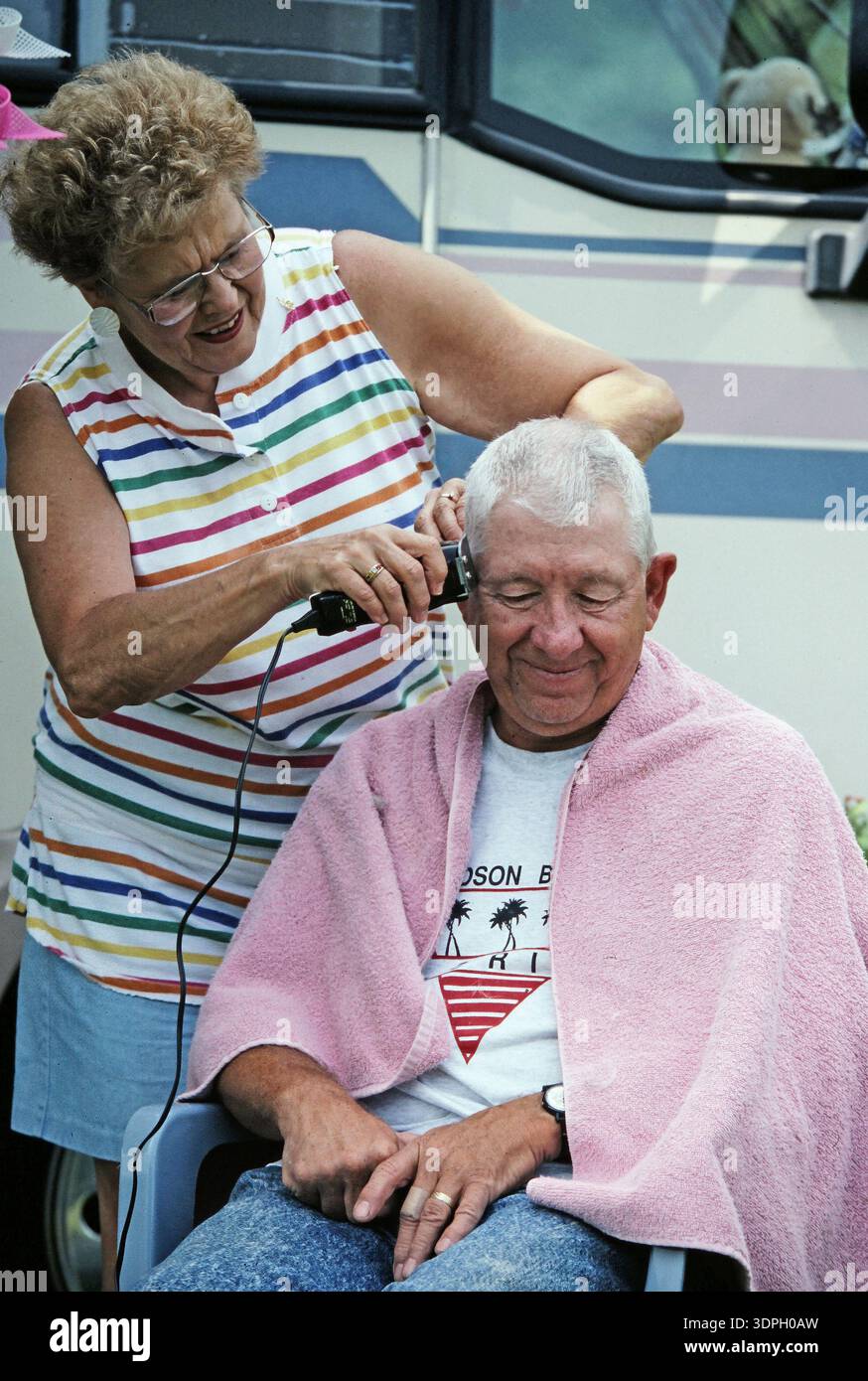 Una donna rifinisce i capelli di un uomo che siede rilassato su una sedia. Sono all'aperto vicino a un camper. L'uomo indossa una camicia leggera e un asciugamano, e la donna lo usa Foto Stock