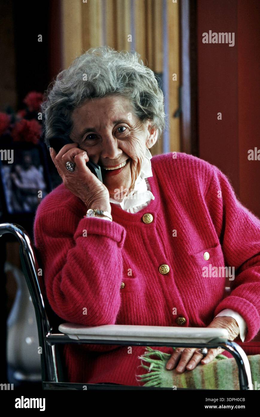 Una donna anziana siede comodamente su una sedia tenendo un telefono all'orecchio. Sorride felicemente, indossando un maglione rosa. L'impostazione ha colori caldi Foto Stock