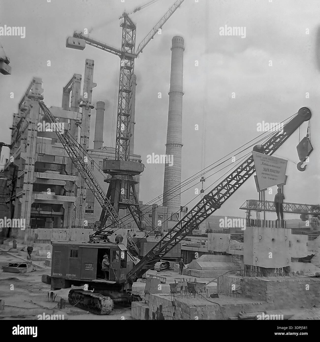 Una notevole fotografia d'archivio in bianco e nero del 1965 che documenta la costruzione su larga scala del blocco di potenza da 800 MW presso la centrale termica Slavyanskaya (GRES) a Nikolaevka, regione di Donetsk. Una gru cingolata pesante è posizionata in primo piano, issando un grande cartello con un impegno socialista per il collettivo Metallomontazh, fissando una scadenza del 15 ottobre 1965. Lo sfondo rivela torreggianti camini e strutture industriali scheletriche. Questo scatto autentico cattura l'immenso sforzo ingegneristico e lo spirito delle pacifiche Donbas. Foto Stock