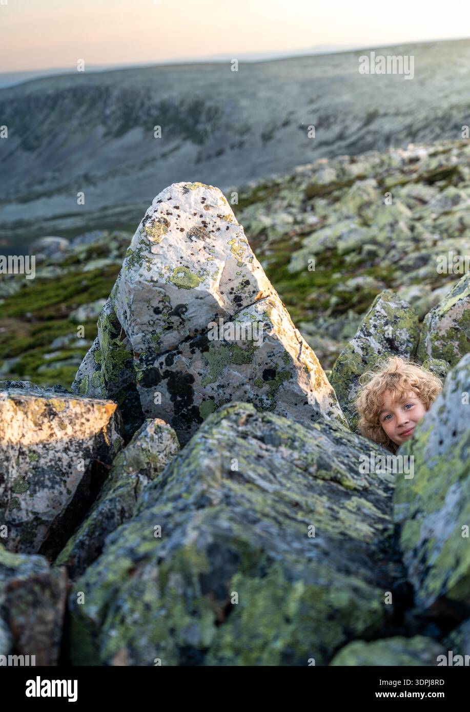 Un ragazzo con i capelli biondi ricci gioca a nascondino tra le grandi rocce di una montagna in Svezia. Sbircia tra i massi guardando la telecamera con un sorriso Foto Stock
