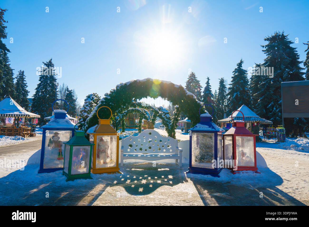 Area fotografica invernale con panchina bianca intagliata, lanterne colorate giganti e archi sempreverdi sotto il sole luminoso in un parco innevato Foto Stock