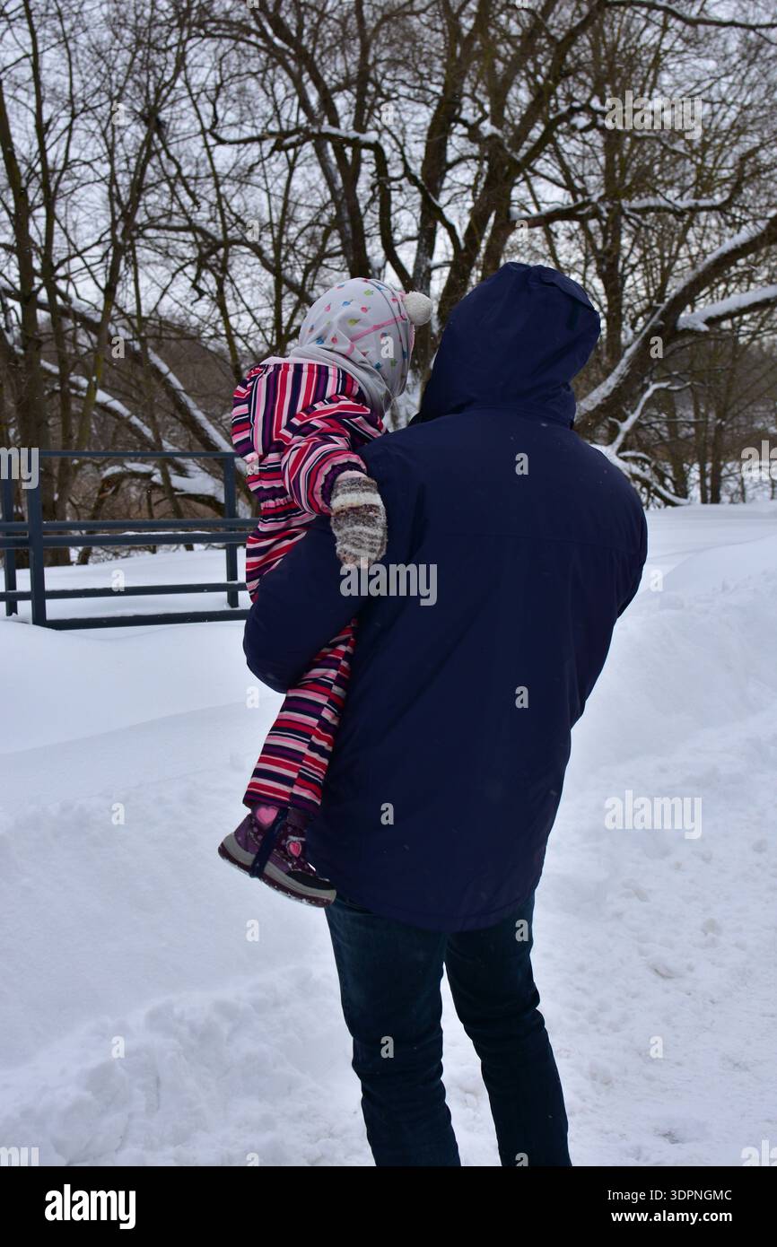 Padre genitore che trasporta una bambina piccola su un sentiero innevato nel parco invernale. Momenti caldi in famiglia all'aperto con il freddo, con alberi innevati e. Foto Stock