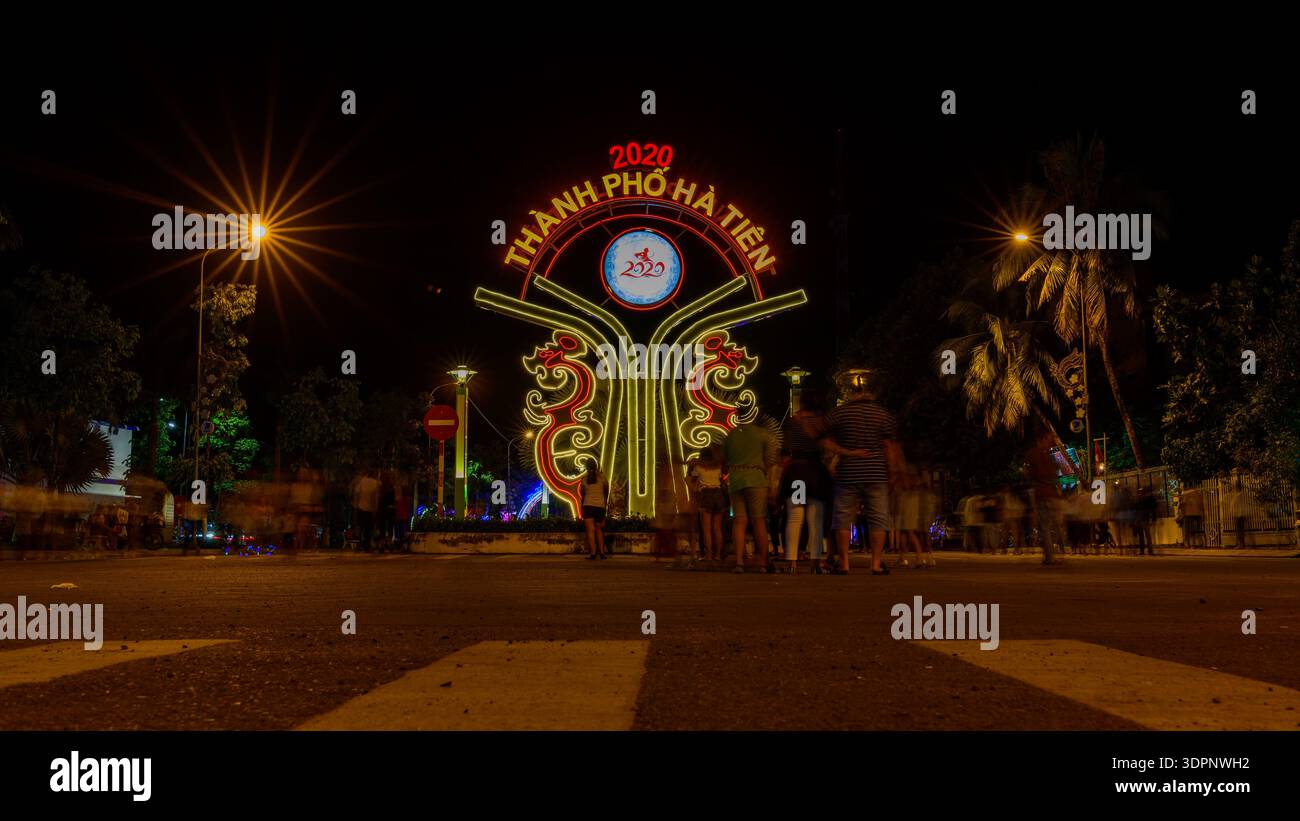 Ha Tien, Vietnam - 24 gennaio 2020: Un gruppo di persone si trova di fronte a un cartello luminoso della porta della città con la scritta "Thanh Pho ha Tien 2020" di notte, con palme, lampioni e alberi visibili sullo sfondo. Foto Stock
