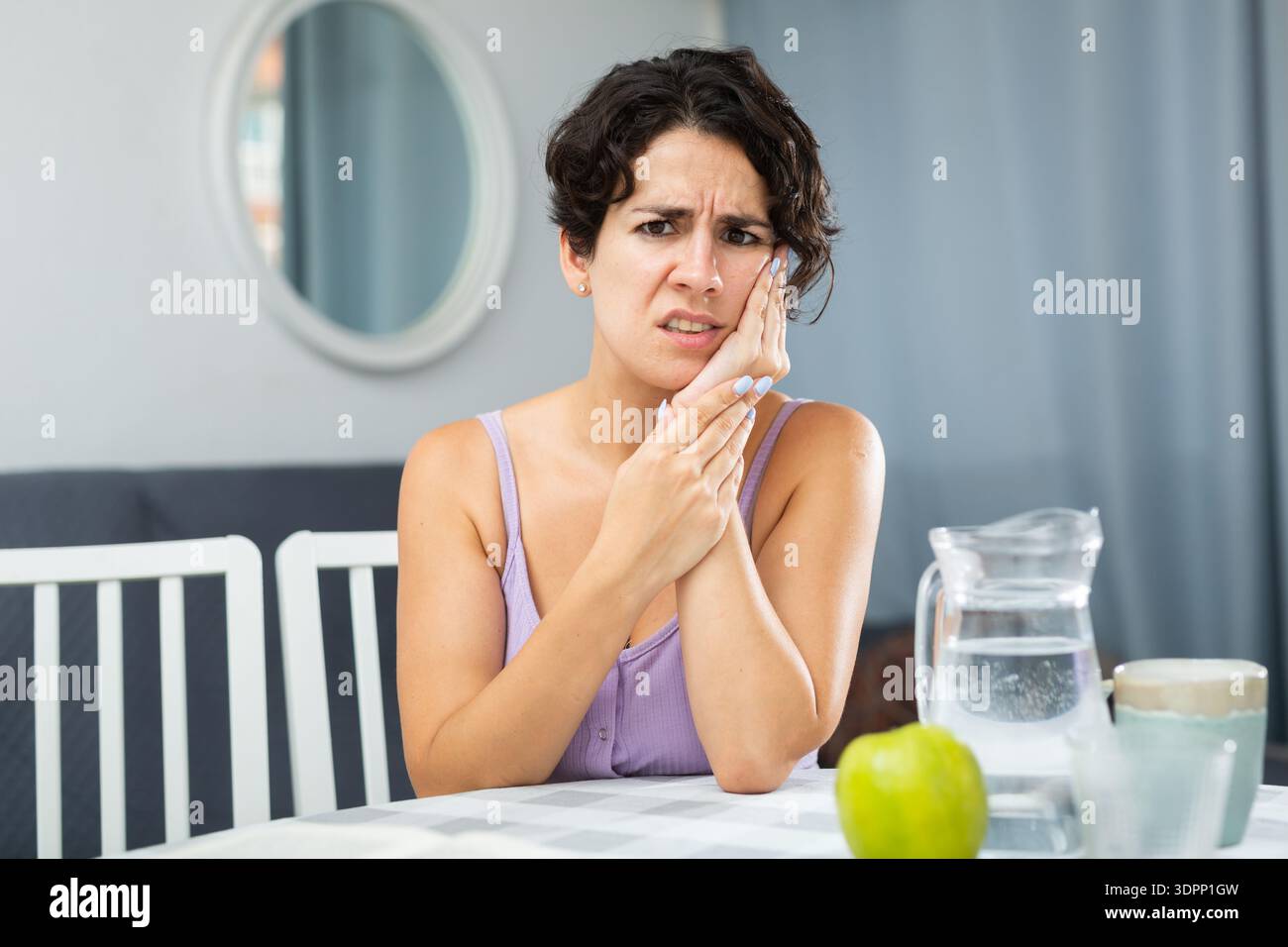 Giovane donna con mal di denti e guancia toccante, seduta a tavola Foto Stock