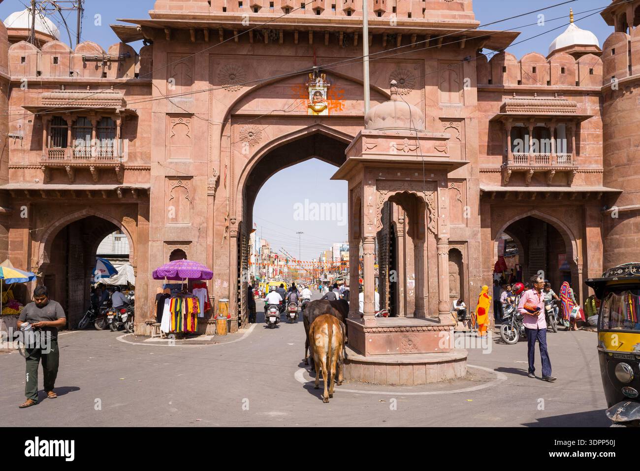 Bikaner, India - 31 marzo 2022: È visibile un grande cancello storico in pietra arenaria con torri a cupola, con persone che camminano, motociclette, risciò auto, una mucca, venditori ambulanti e bancarelle intorno all'ingresso sotto un cielo limpido. Foto Stock