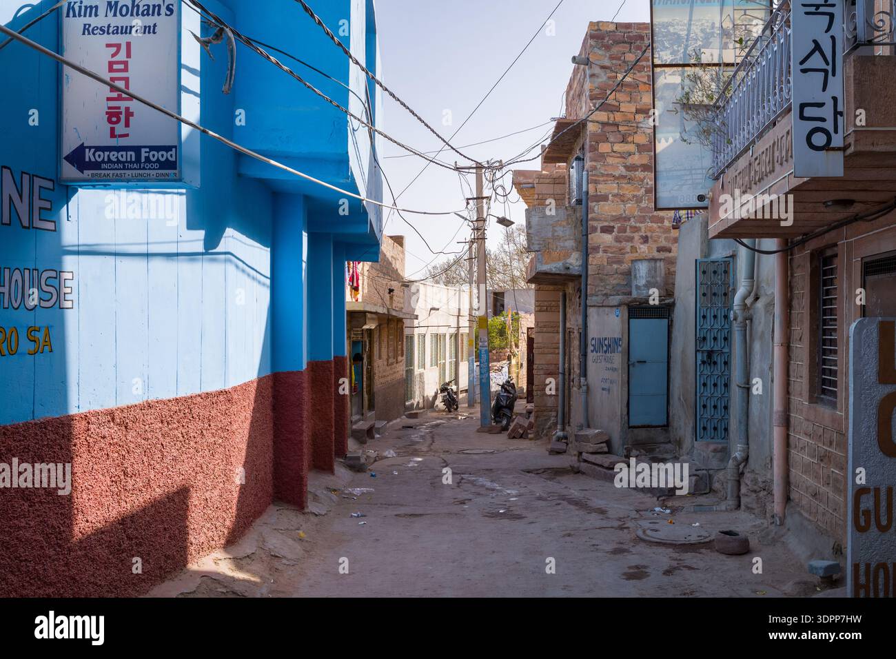 Jodhpur, India - 26 marzo 2022: Uno stretto vicolo è fiancheggiato da edifici blu e in pietra, cartellonistica coreana visibile in inglese e coreano, cavi elettrici sospesi e un sentiero di terra sotto la luce del giorno. Foto Stock