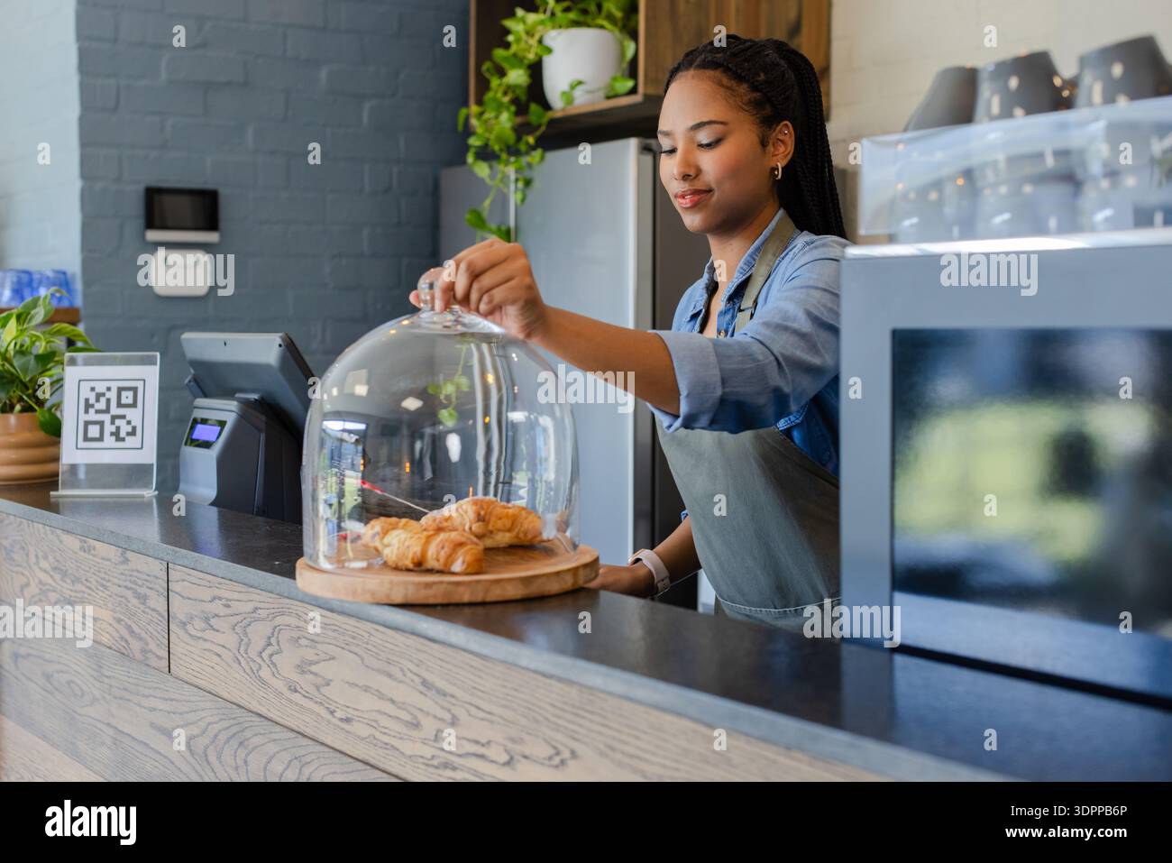 Donna afroamericana in grembiule che solleva la cupola di vetro sul bancone del caffè che mostra croissant presso il sistema POS Foto Stock