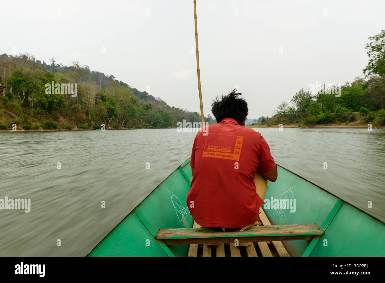 Villaggio degli elefanti, Laos - 25 marzo 2020: Un uomo che indossa una camicia rossa con testo sul retro si siede alla prua di una barca di legno verde che si muove lungo un fiume, con colline boscose e alberi visibili su entrambe le rive del fiume. Foto Stock