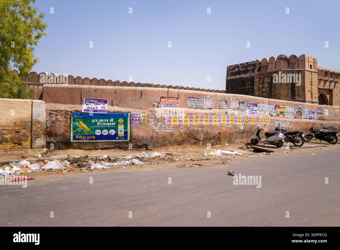 Bikaner, India - 31 marzo 2022: Una scena di strada mostra uno storico muro di fortificazione coperto da poster e pubblicità, con diverse motociclette parcheggiate e rifiuti sparsi visibili lungo la strada sotto un cielo limpido. Foto Stock