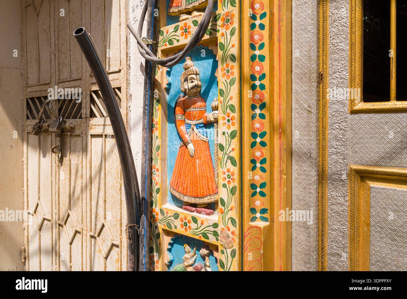 Udaipur, India - 23 marzo 2022: Vista ravvicinata di una porta in legno intagliato e dipinto con una tradizionale figura Rajasthani in abbigliamento arancione, motivi floreali e porte metalliche adiacenti, con cavi elettrici visibili alla luce del sole. Foto Stock