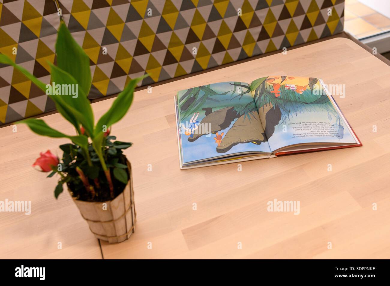 Luogo sconosciuto - 12 maggio 2021: Un libro illustrato aperto per bambini è collocato su un tavolo di legno chiaro accanto ad una pianta in vaso con un fiore rosa, con una panchina geometrica sullo sfondo. Foto Stock