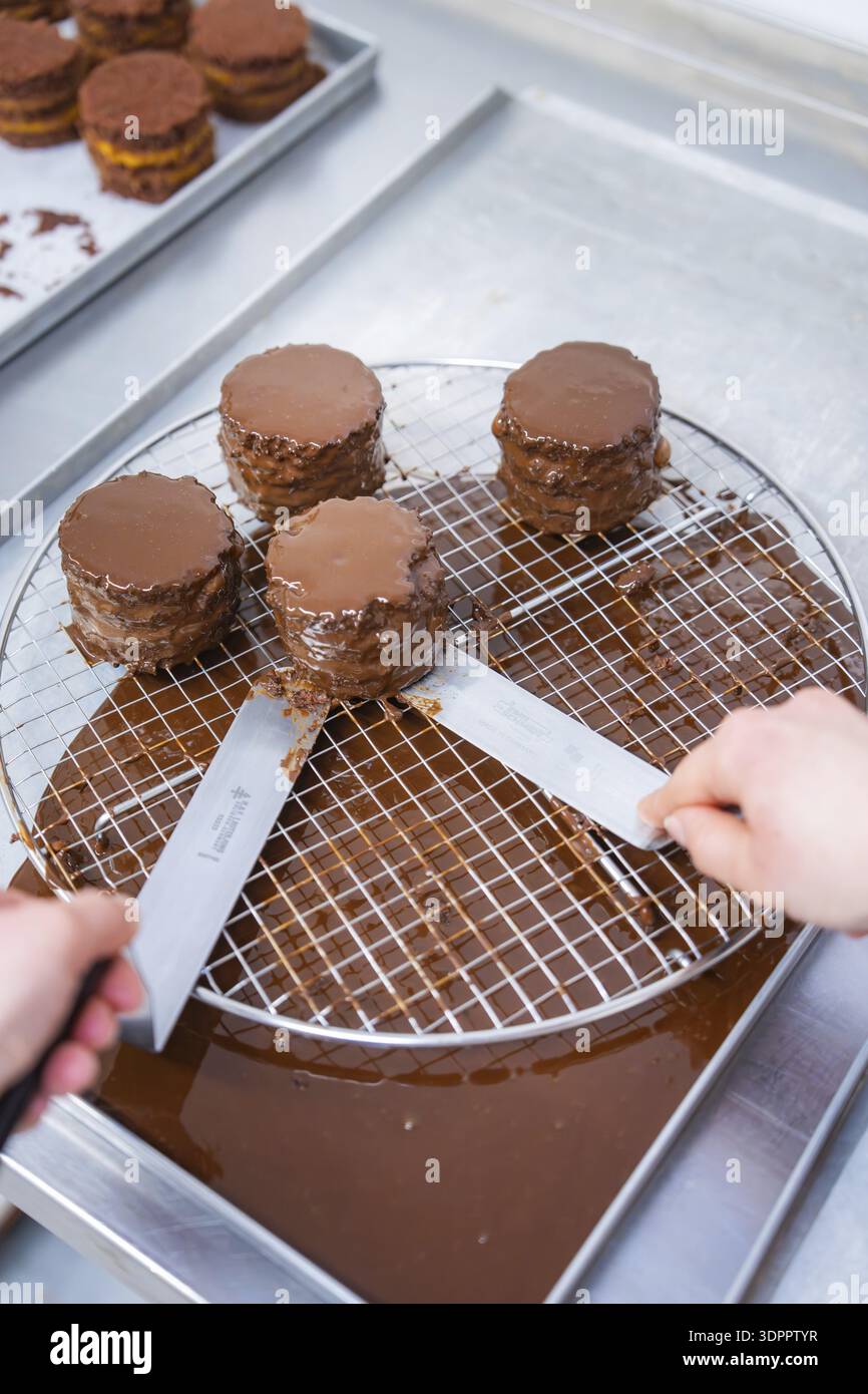 Due mani usano i coltelli per spostare diverse torte glassate al cioccolato su una rete gocciolante in cucina, produzione di torte vegane, Haselstaller Hof, Gechingen, Germania Foto Stock