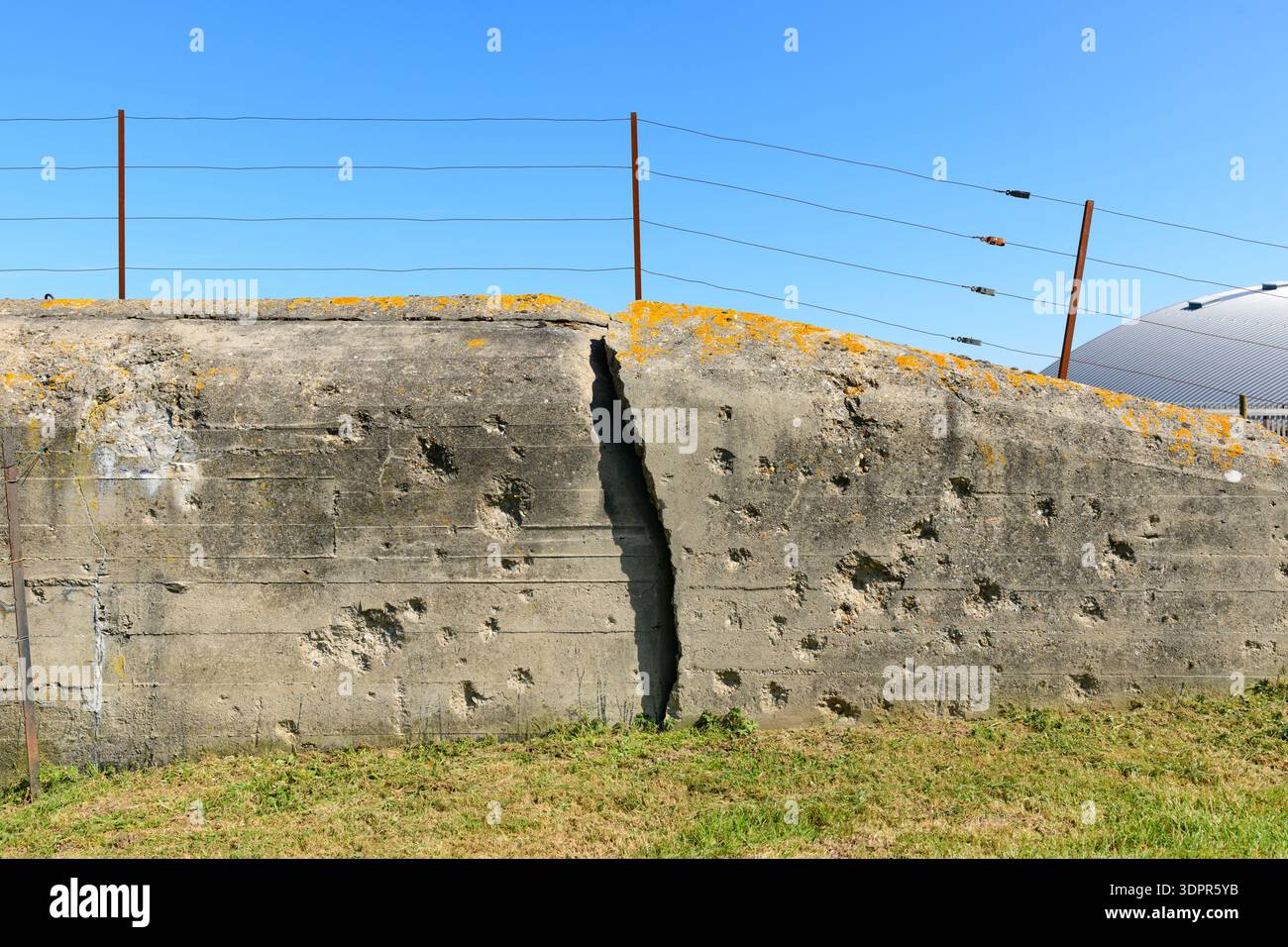 Batterie de Merville, Francia - 13 aprile 2021: Vista ravvicinata di un muro di cemento incrinato e danneggiato dell'era della seconda guerra mondiale con segni di impatto visibili, sormontato da recinzioni a filo metallico e lichene giallo a batterie de Merville. Foto Stock