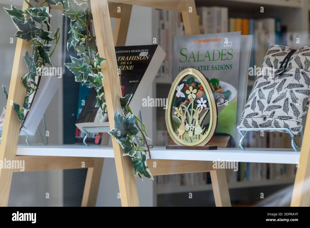 Luogo sconosciuto - 12 maggio 2021: Libri con titoli francesi visibili, un uovo decorativo in legno con motivi floreali e conigli, e una custodia in tessuto a motivi geometrici sono disposti su una mensola bianca dietro il vetro, con edera artificiale drappeggiata su una cornice in legno. Foto Stock