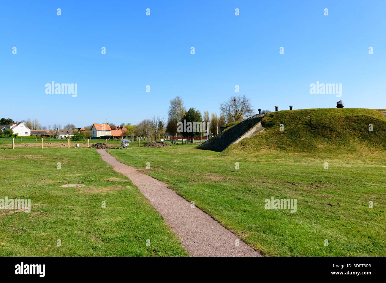 Batterie de Merville, Francia - 13 aprile 2021: Tumulo erboso con strutture di ventilazione e una scalinata in cemento, con un sentiero e case residenziali visibili sullo sfondo a batterie de Merville. Foto Stock