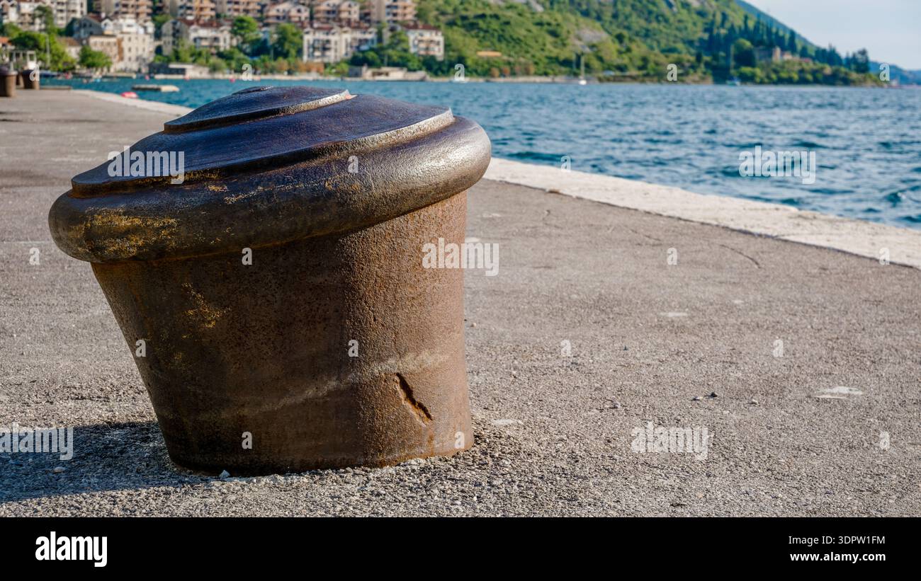 Un dissuasore metallico intemprato si trova su un molo di cemento vicino all'acqua. Lo sfondo presenta verdi colline e una città lontana lungo il litorale. Foto Stock