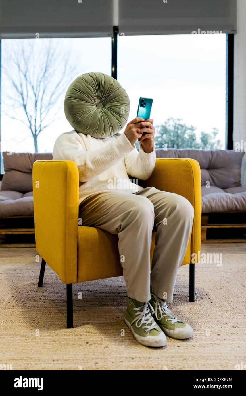 Una persona con un cuscino che copre la testa è seduta su una poltrona gialla, utilizzando uno smartphone. Gli interni accoglienti dispongono di un comodo divano e di una creatina Foto Stock