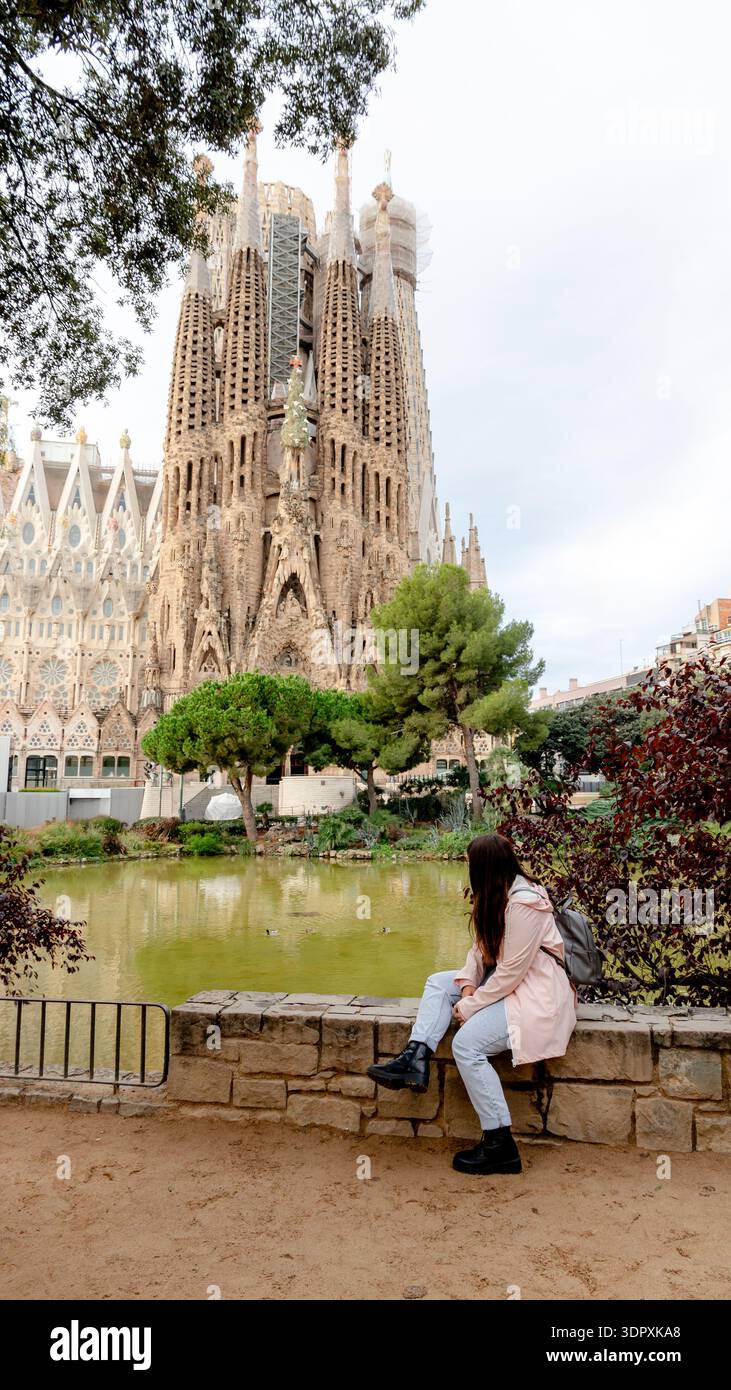 Una donna siede su una sporgenza di pietra, osservando attentamente l'iconica Sagrada Familia di Barcellona. Questo capolavoro architettonico di Antoni Gaudí continua Foto Stock